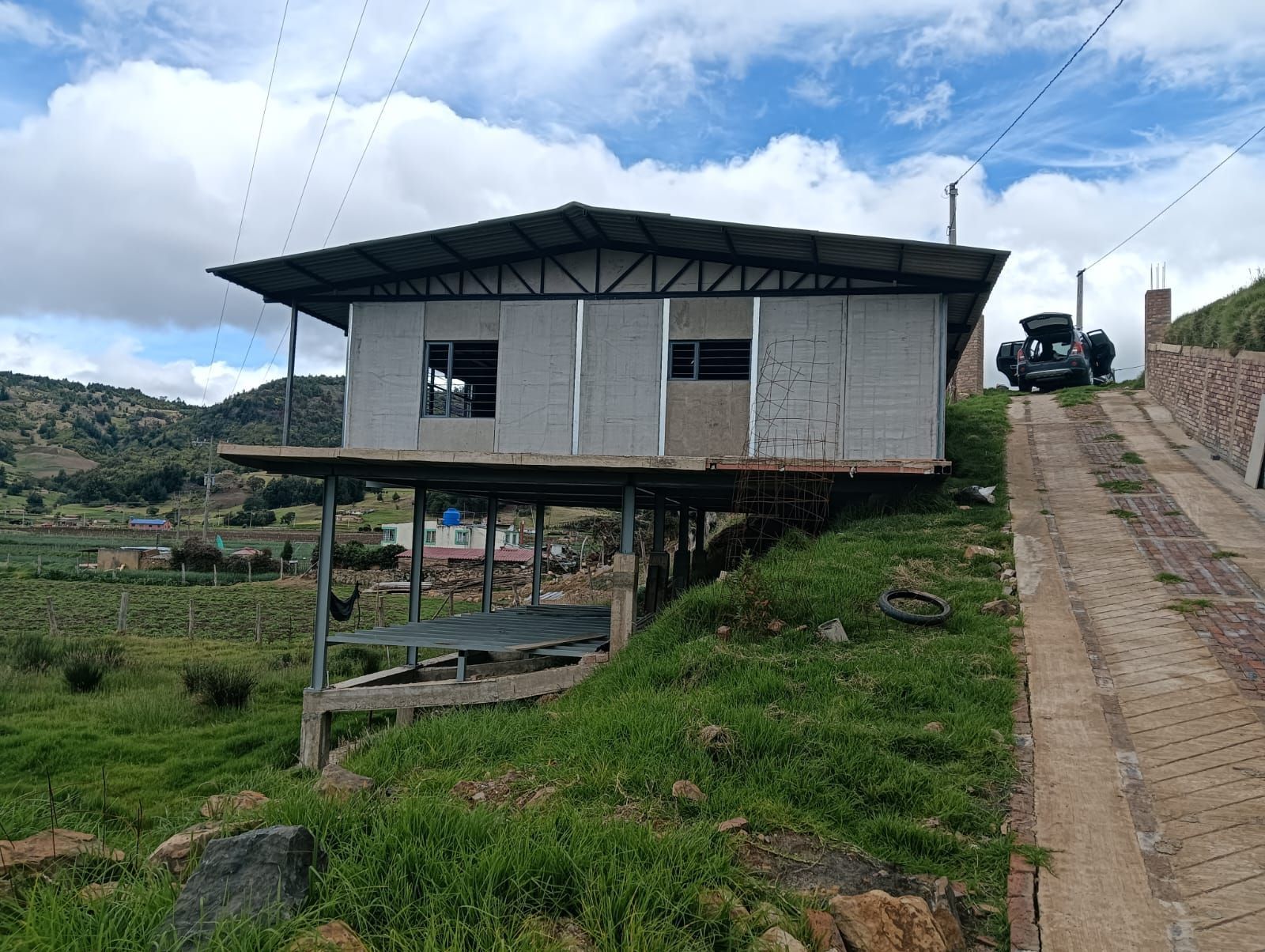 Pequeña casa elevada sobre pilotes junto a una carretera empinada en un pueblo de ladera cubierta de hierba.