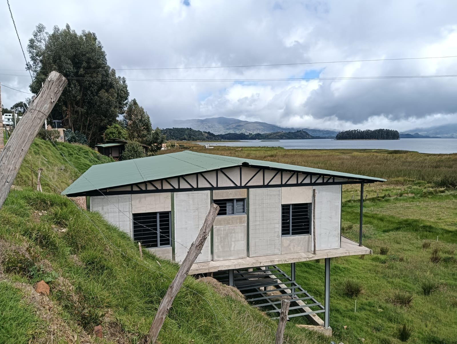 Pequeña casa blanca con techo verde en una ladera cubierta de hierba junto al agua y las montañas bajo un cielo nublado.