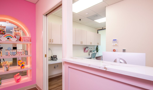 A bright pink office reception area with a white marble counter, open shelving with rainbow decor, and cabinets.