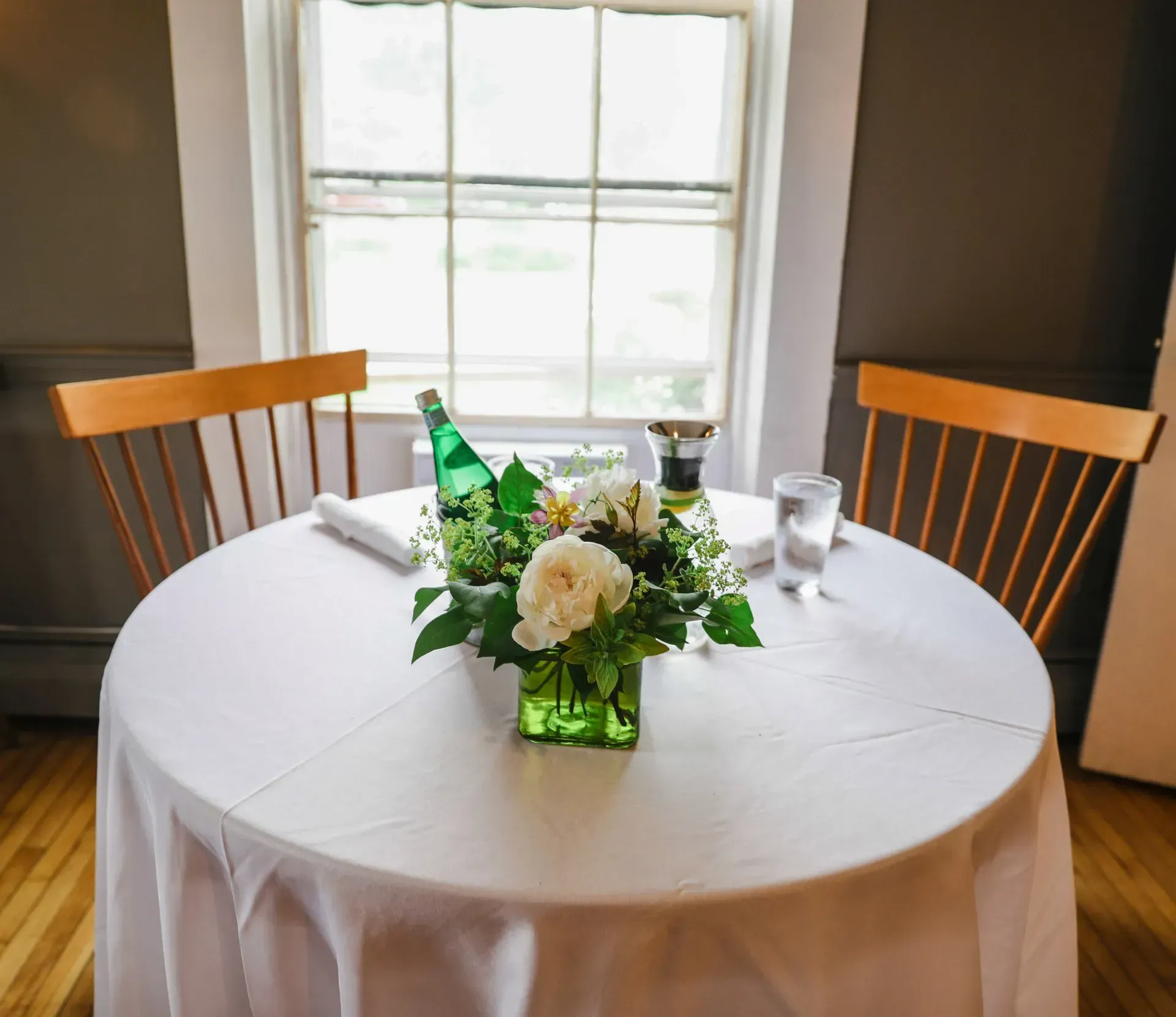 Small round table with white cloth, floral centerpiece, and two wooden chairs by a window