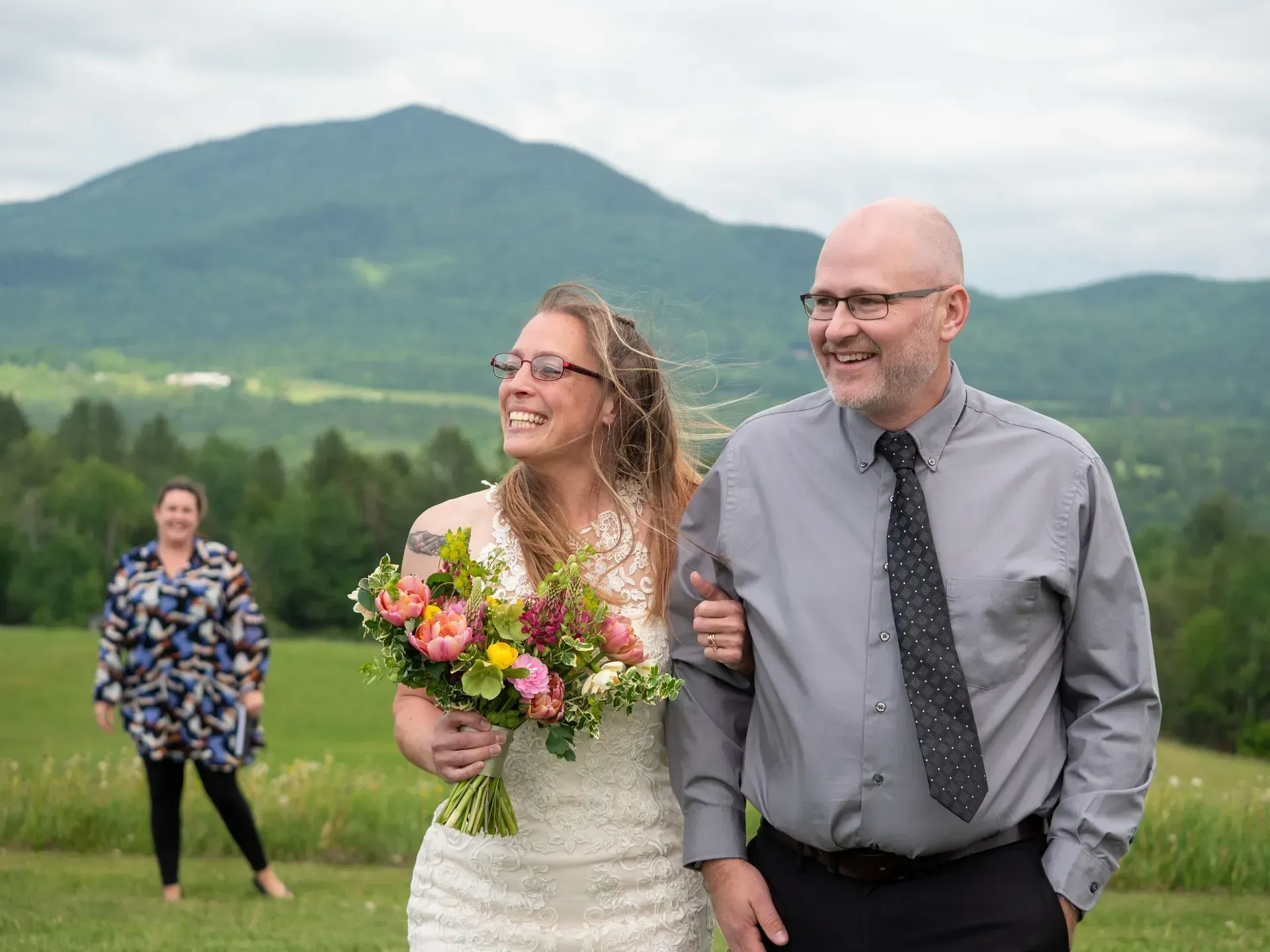 Smiling couple outdoors with a bouquet, walking in front of a green mountain landscape