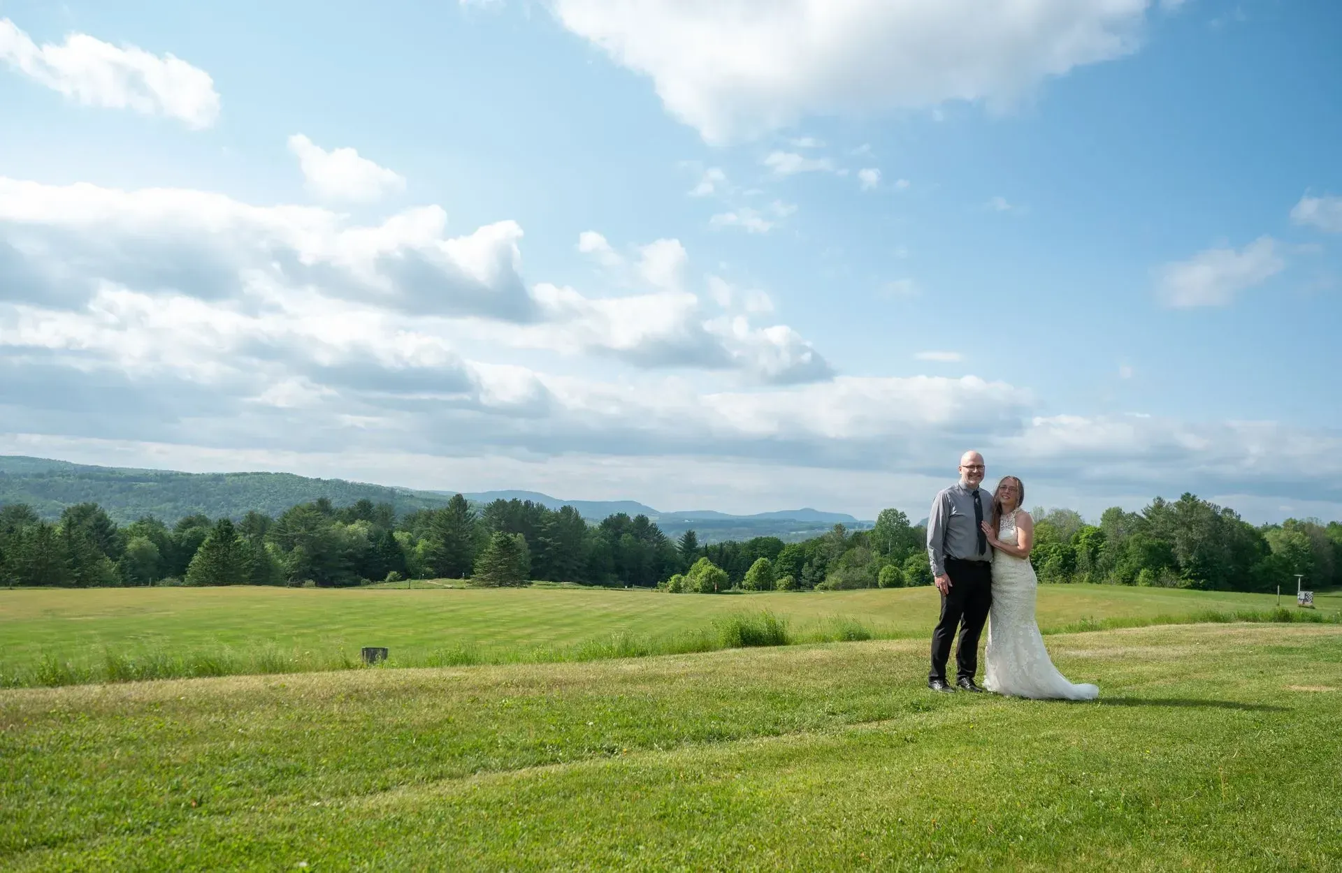 Bride and groom standing on a grassy field under a blue sky with clouds