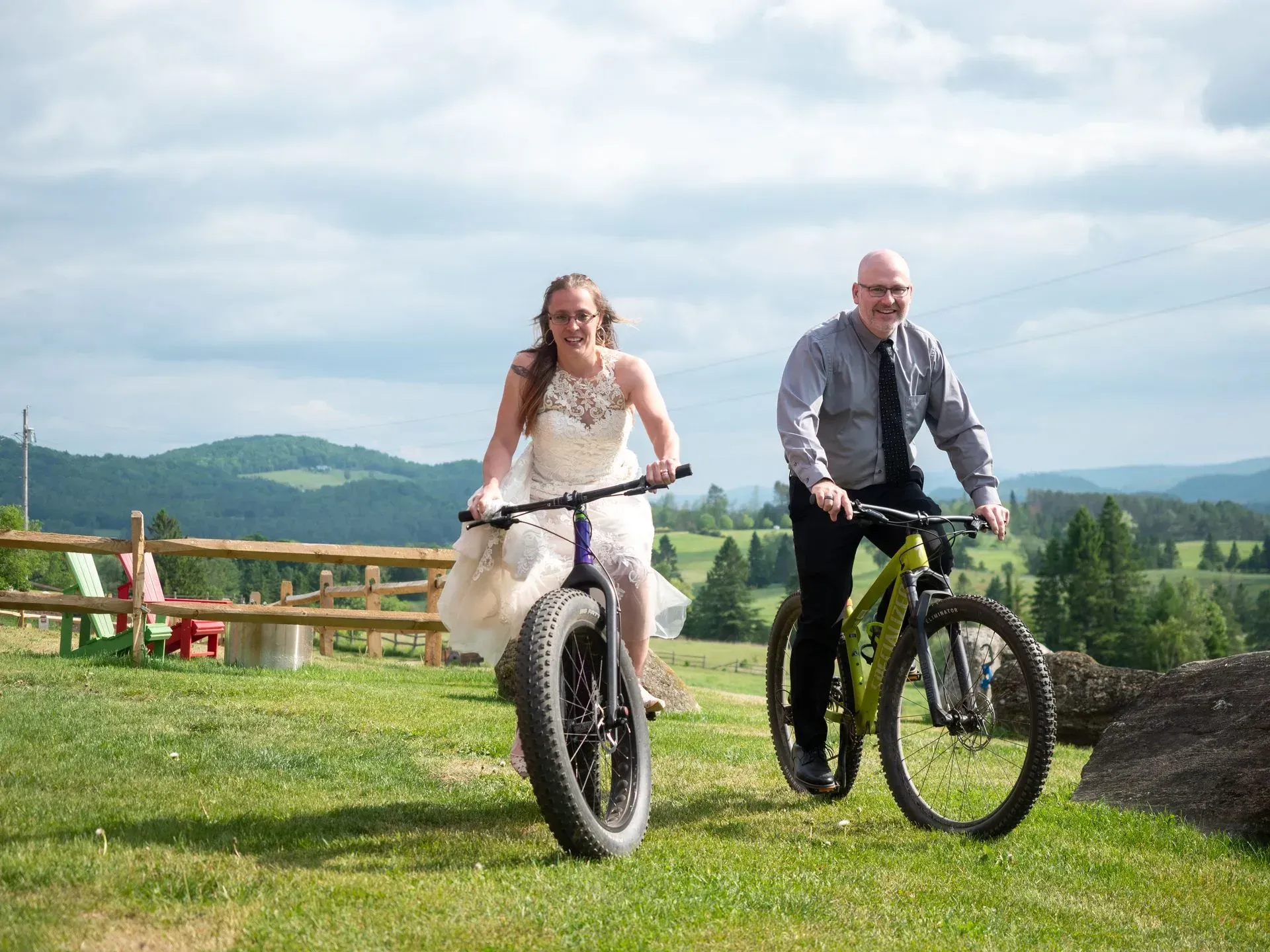 Two people riding bicycles on a grassy hill with mountains and blue sky in the background