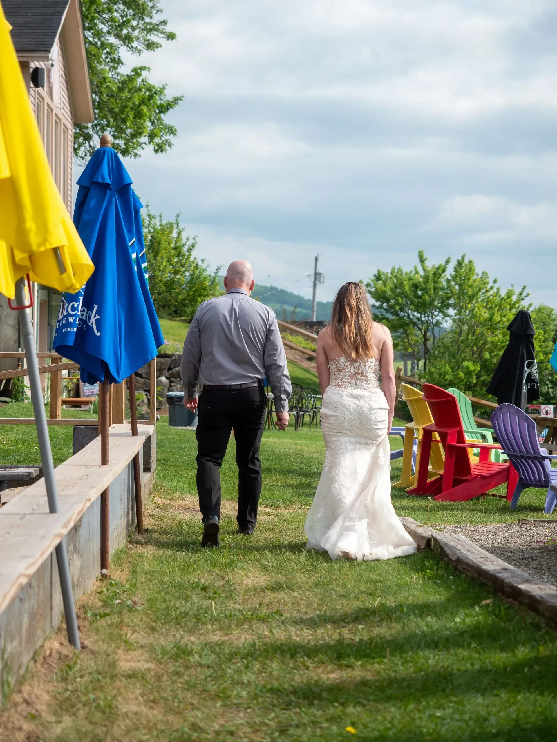 Couple walking down a grassy path at an outdoor wedding with colorful chairs and umbrellas