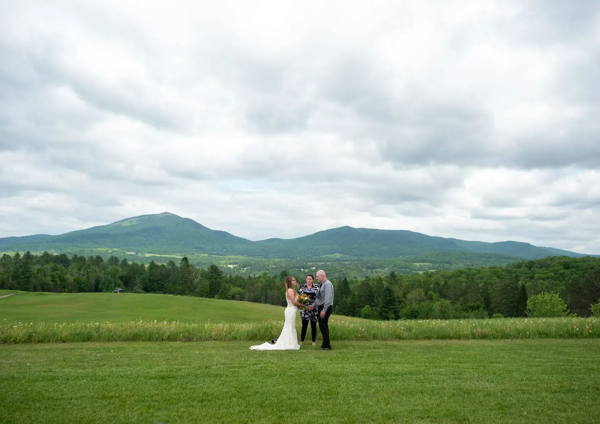 Bride and groom stand in a green field with mountains and cloudy sky behind them.