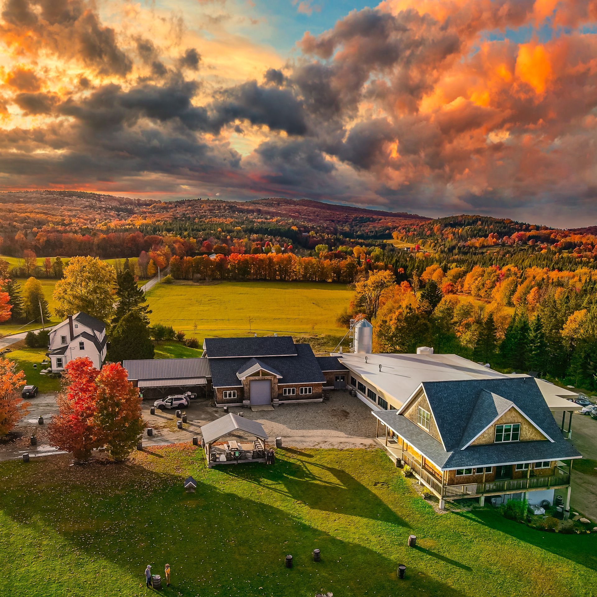 Aerial view of a Hill Farmstead Brewing's farmhouse and barns beside autumn fields under dramatic orange clouds.