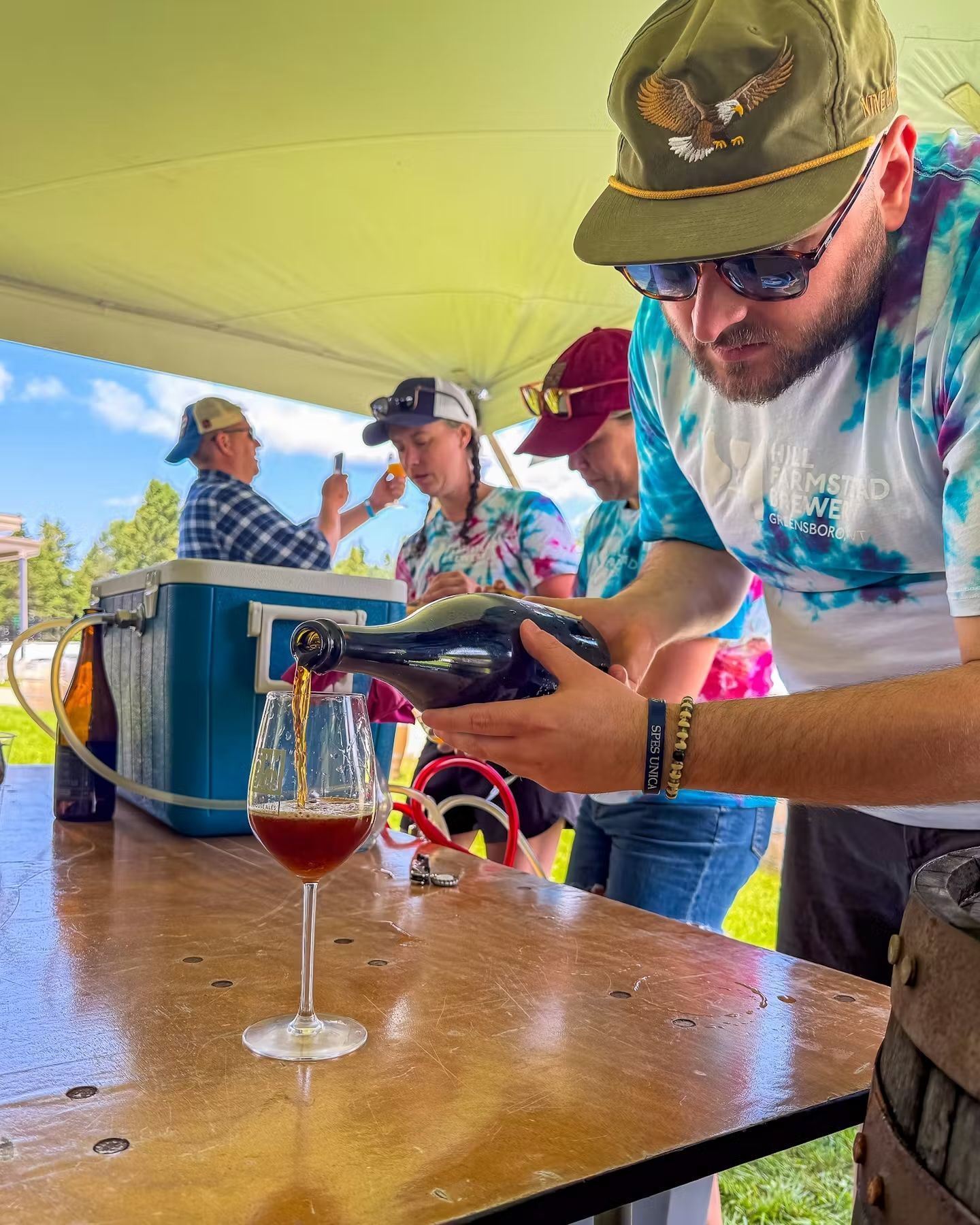 Man pouring Hill Farmstead Brewing's ale into a glass at an outdoor event under a yellow canopy