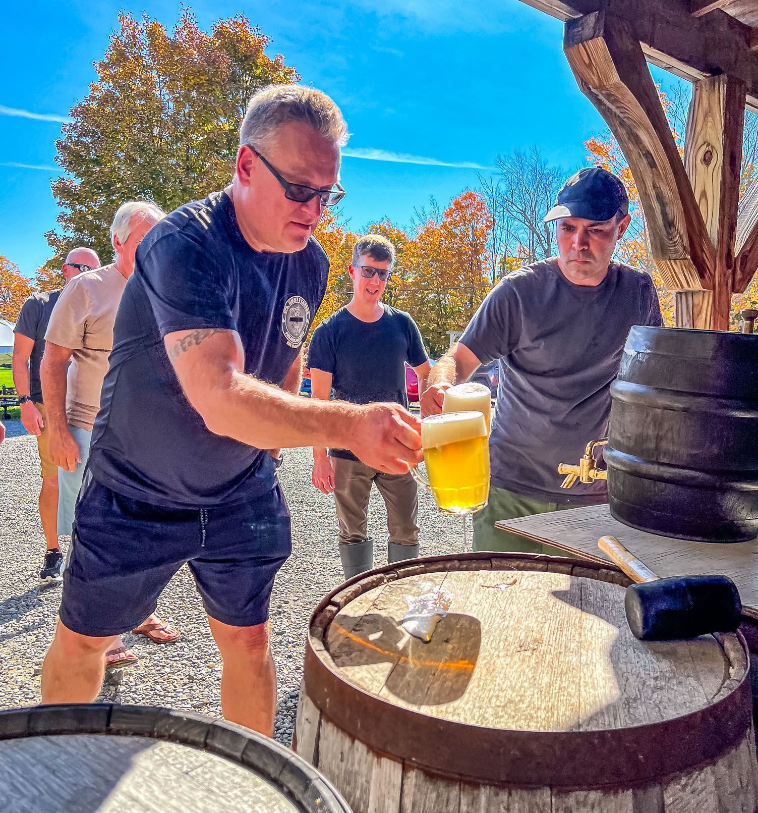 Person pours beer into a glass at an outdoor brewery, with others watching nearby.