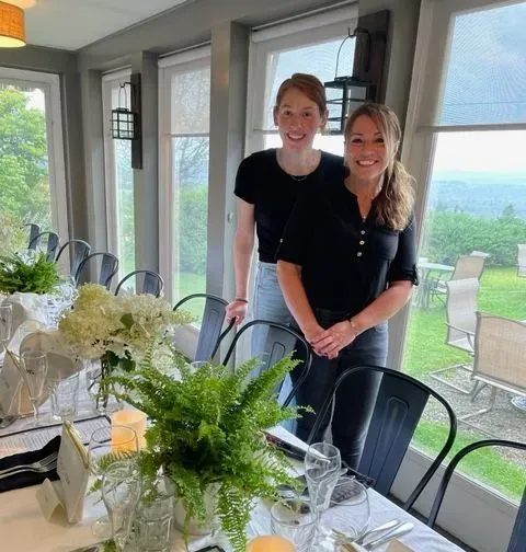 Two smiling people standing beside a decorated dining table in a bright room with large windows and greenery outside