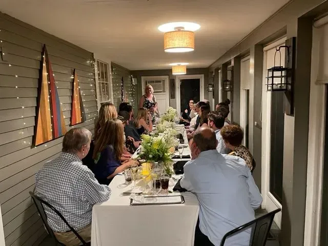 Outdoor dinner party on a covered patio, with guests seated around a long table lit by warm overhead lights.