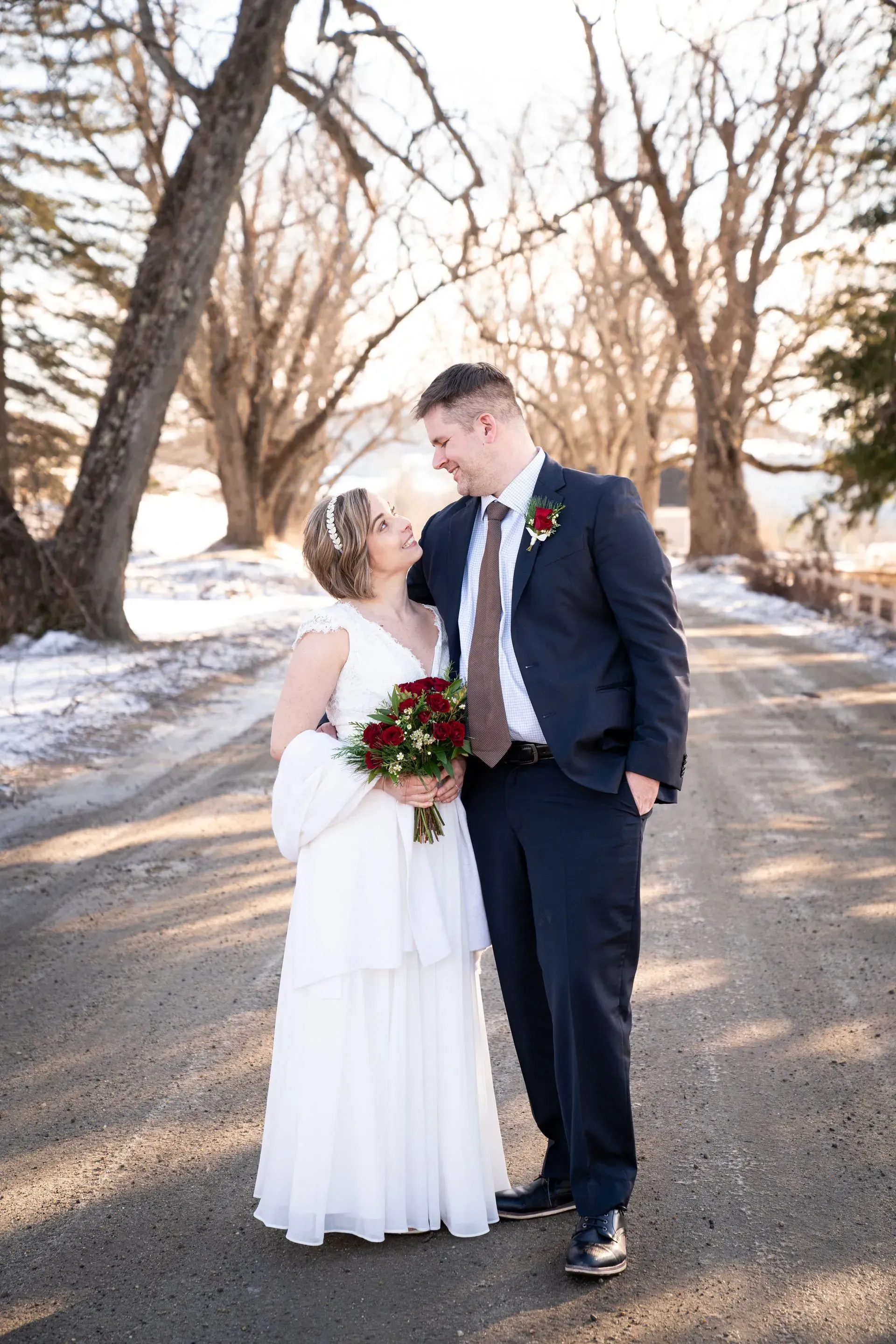Bride and groom standing on a snowy tree-lined road, smiling at each other, holding a bouquet