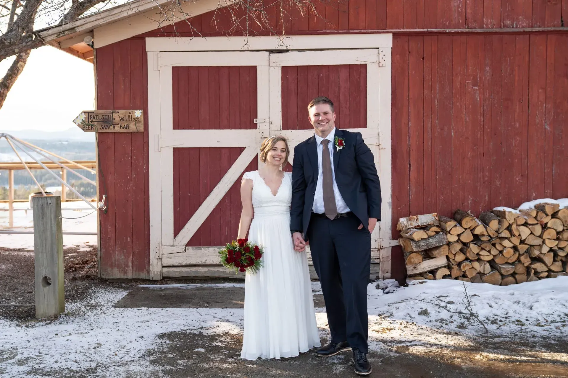 Newlyweds posing outside a red barn in winter, bride in white dress and groom in dark suit.