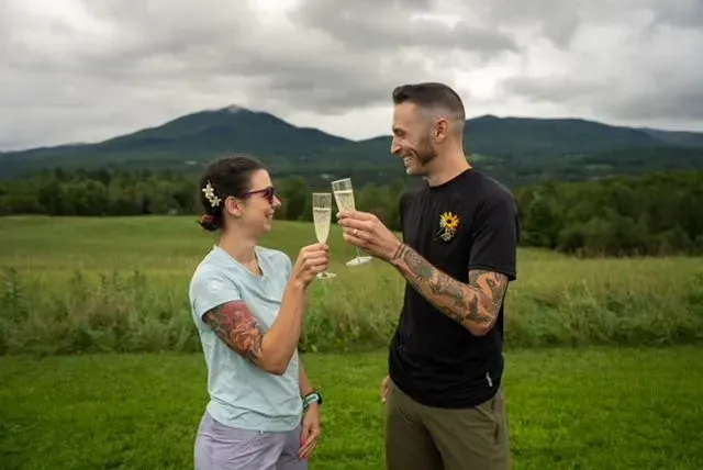Two people clink champagne glasses in a grassy field with mountains in the background.