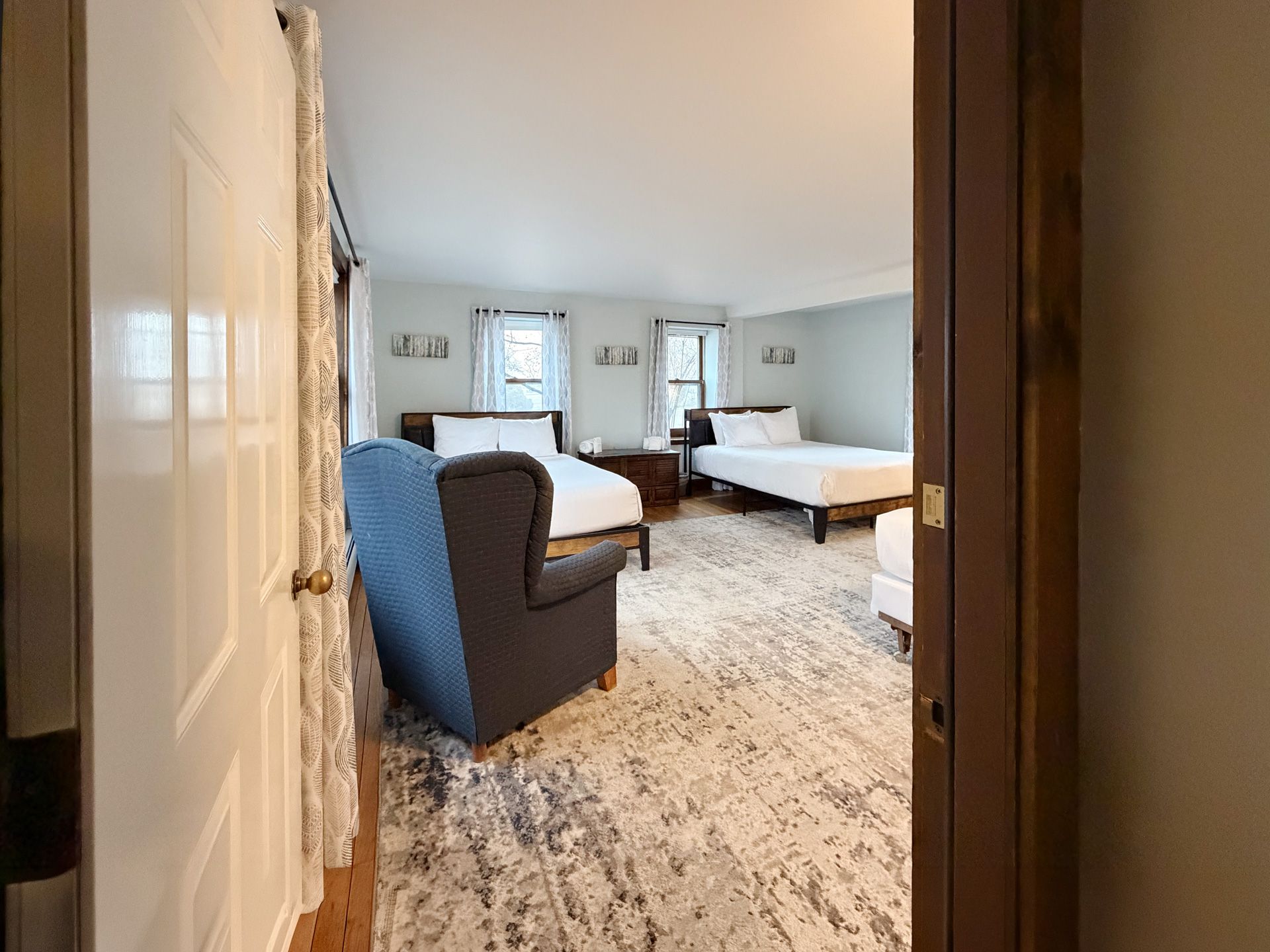Bright bedroom with two white beds, a blue chair, and a light patterned rug viewed through a doorway