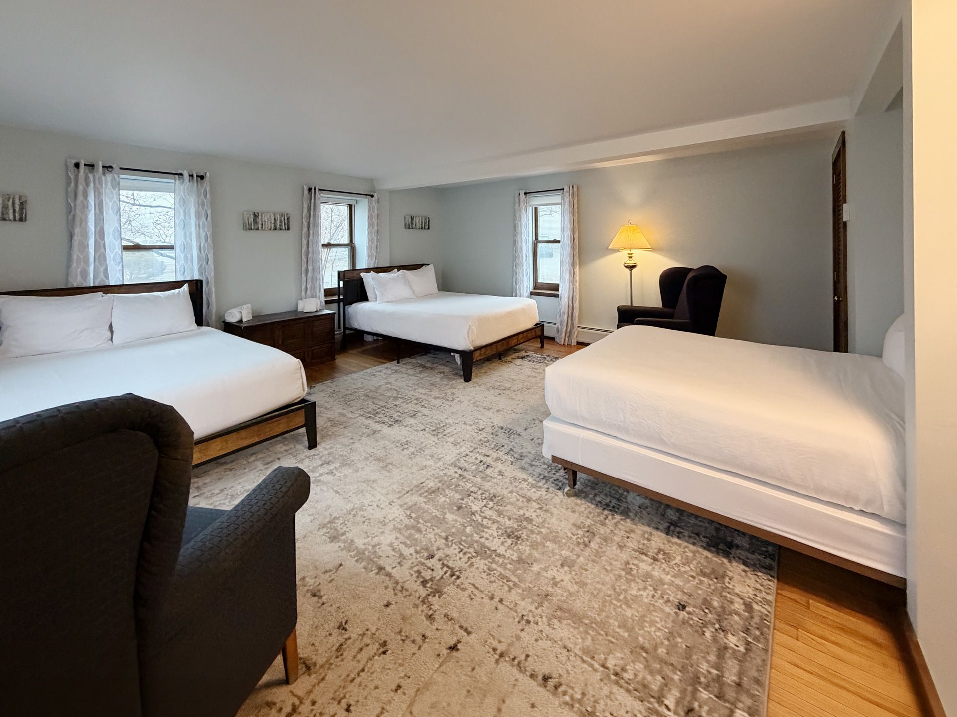 Spacious bedroom with three twin beds, a chair, lamps, and a patterned rug in a light gray room