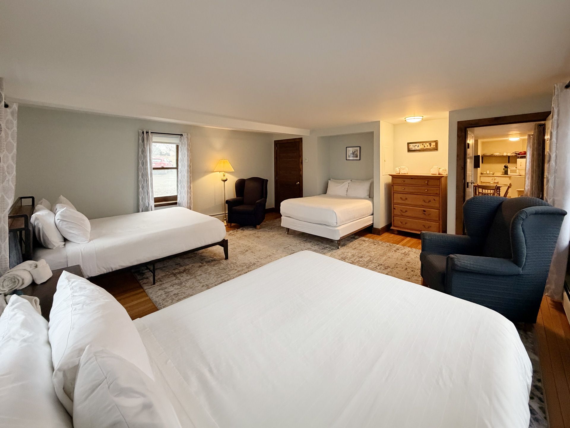 Spacious bedroom with three white beds, gray walls, a blue armchair, and a wooden dresser under warm lighting