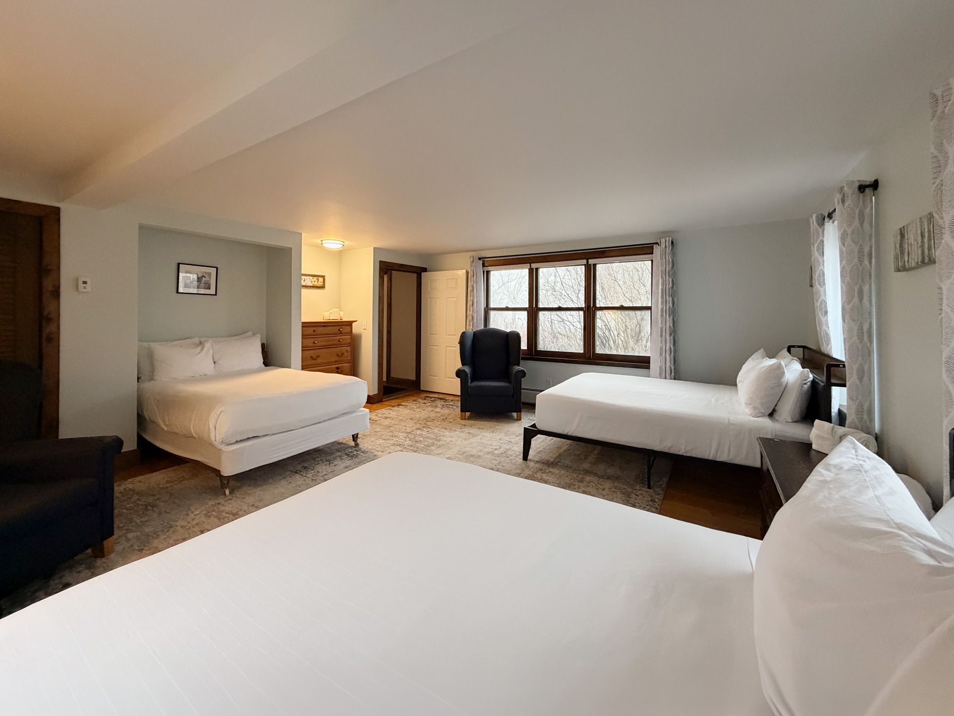 Spacious bedroom with three white beds, a dark armchair, and a bay window with curtains