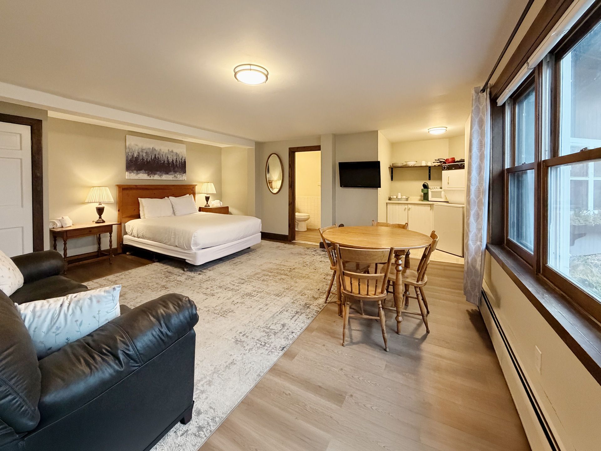 Studio bedroom with queen bed, sofa, dining table, and large windows in warm neutral tones