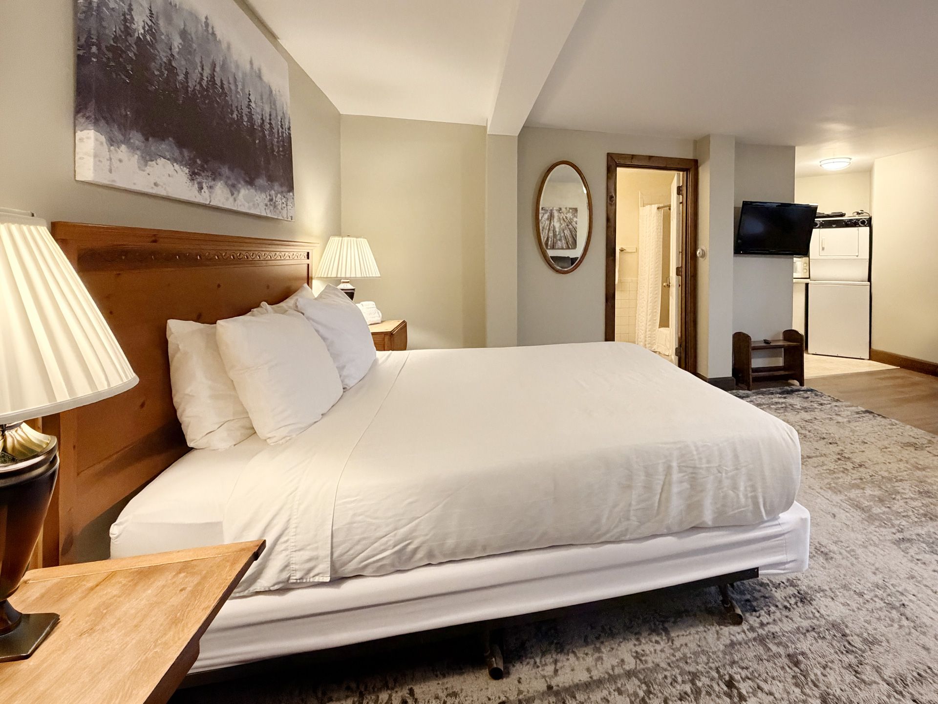 Hotel bedroom with a large white bed, wooden headboard, bedside lamps, and TV in the background