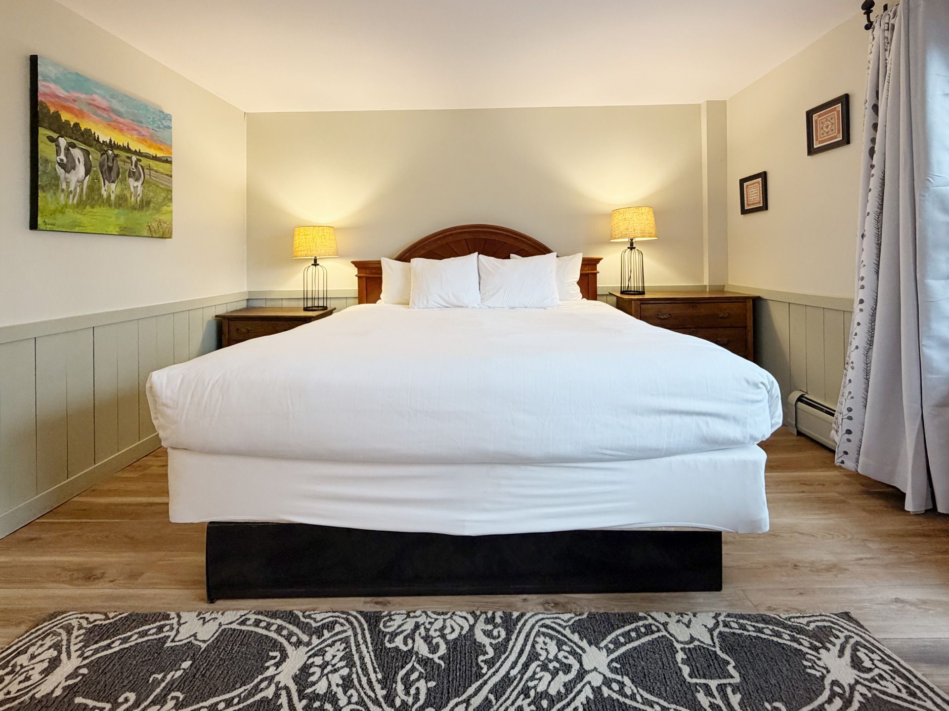 Bright bedroom with a large bed, bedside lamps, wall art, and curtains over a patterned rug.