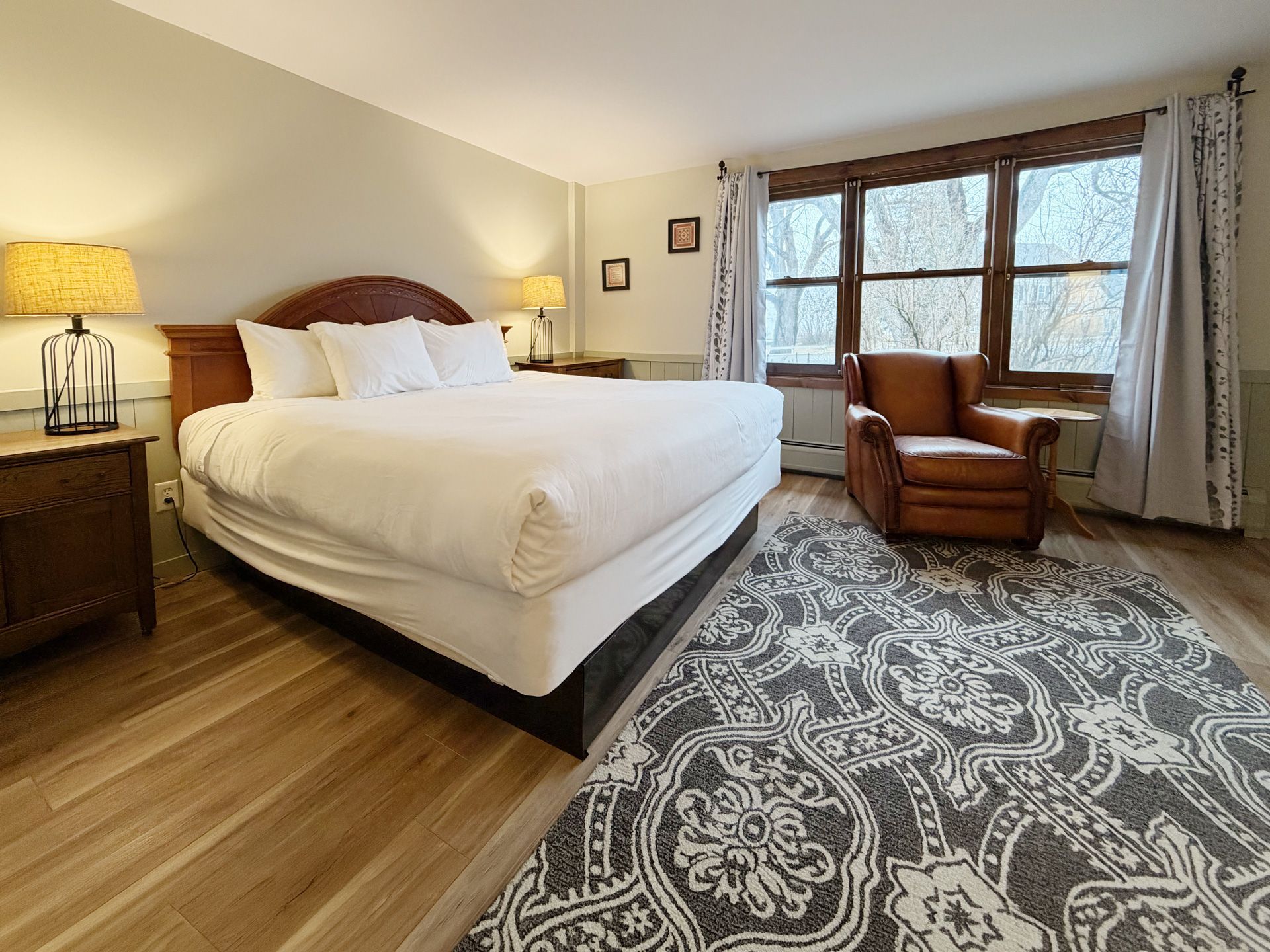 Cozy bedroom with a large bed, wooden nightstand, patterned rug, and window seat by tall windows