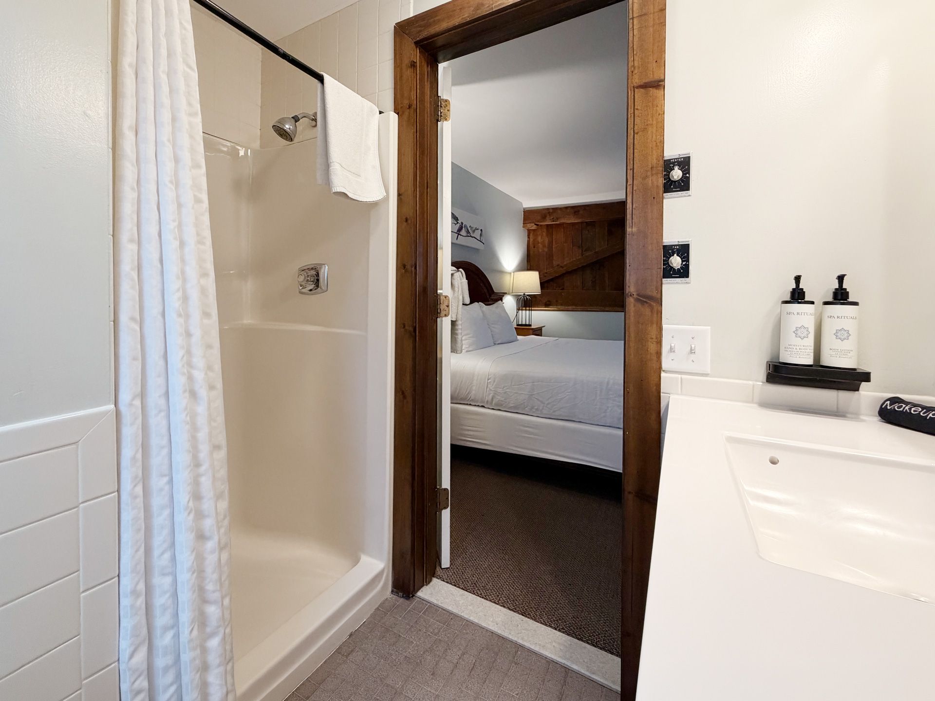 Bathroom doorway with a shower curtain, vanity, and view into a neatly made bed in the adjoining bedroom