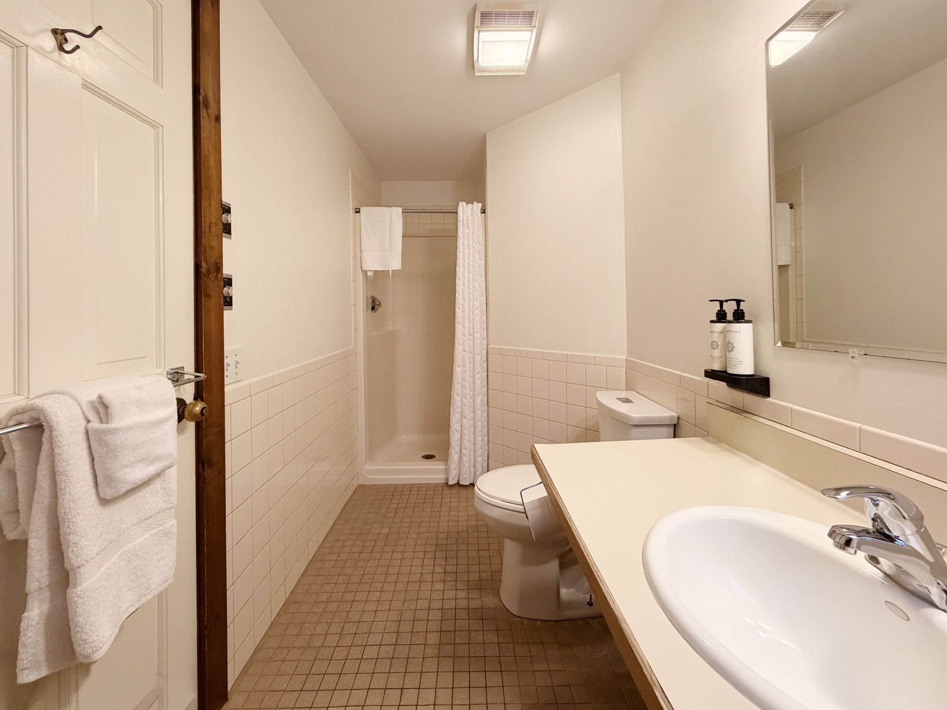 Narrow beige bathroom with sink, mirror, toilet, shower, and wall-mounted towel rack.