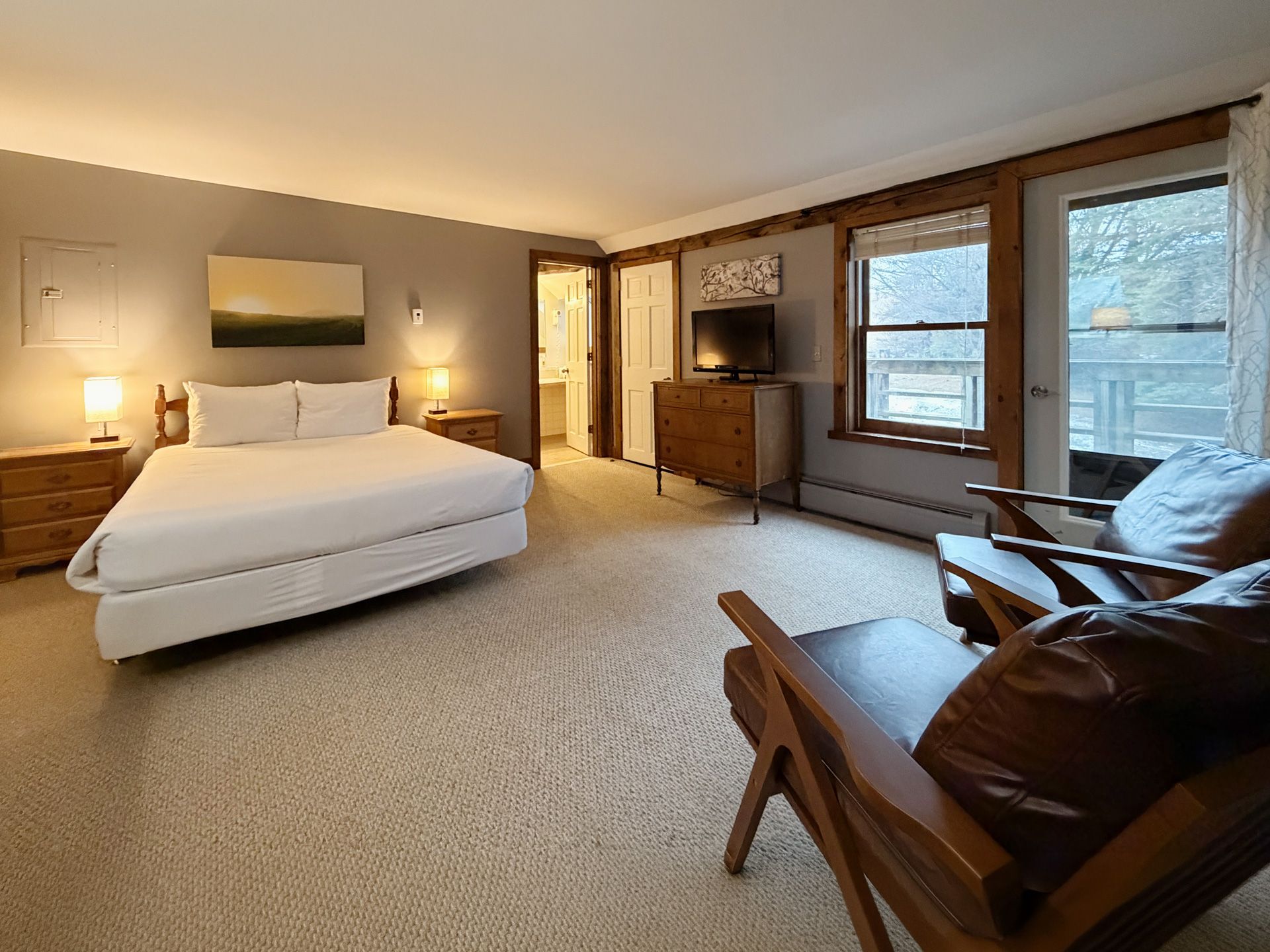 Bright bedroom with a made bed, wood furniture, tan carpet, and windows overlooking snowy trees