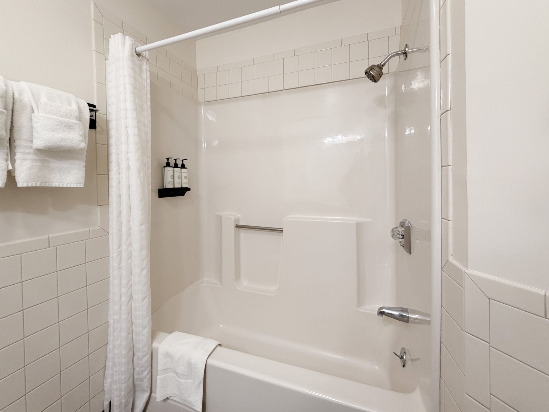 White tiled bathroom with a shower-tub, curtain, grab bar, and towels hanging nearby