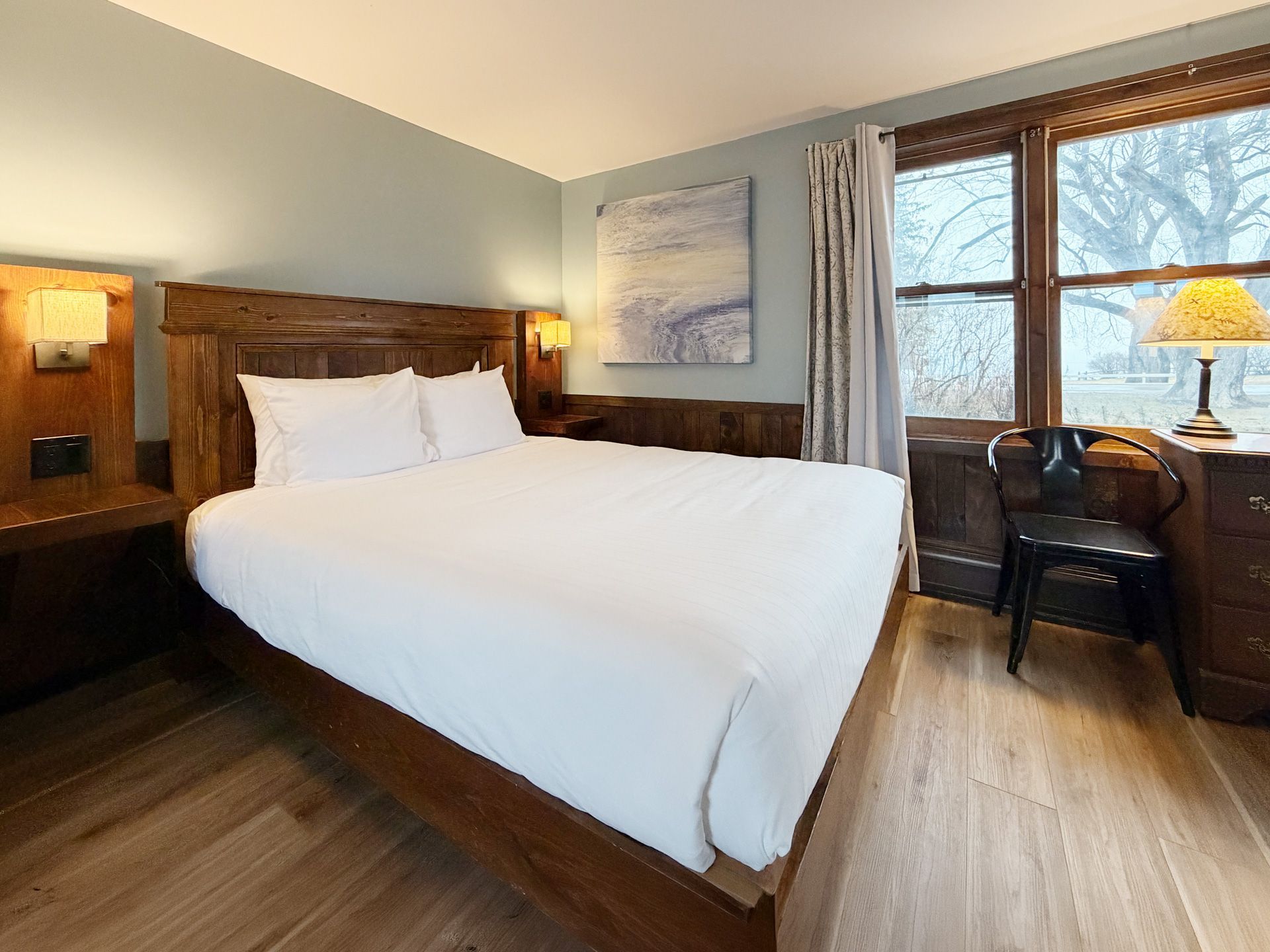 Cozy hotel room with a wooden bed, white bedding, desk, chair, and large window with a lamp.