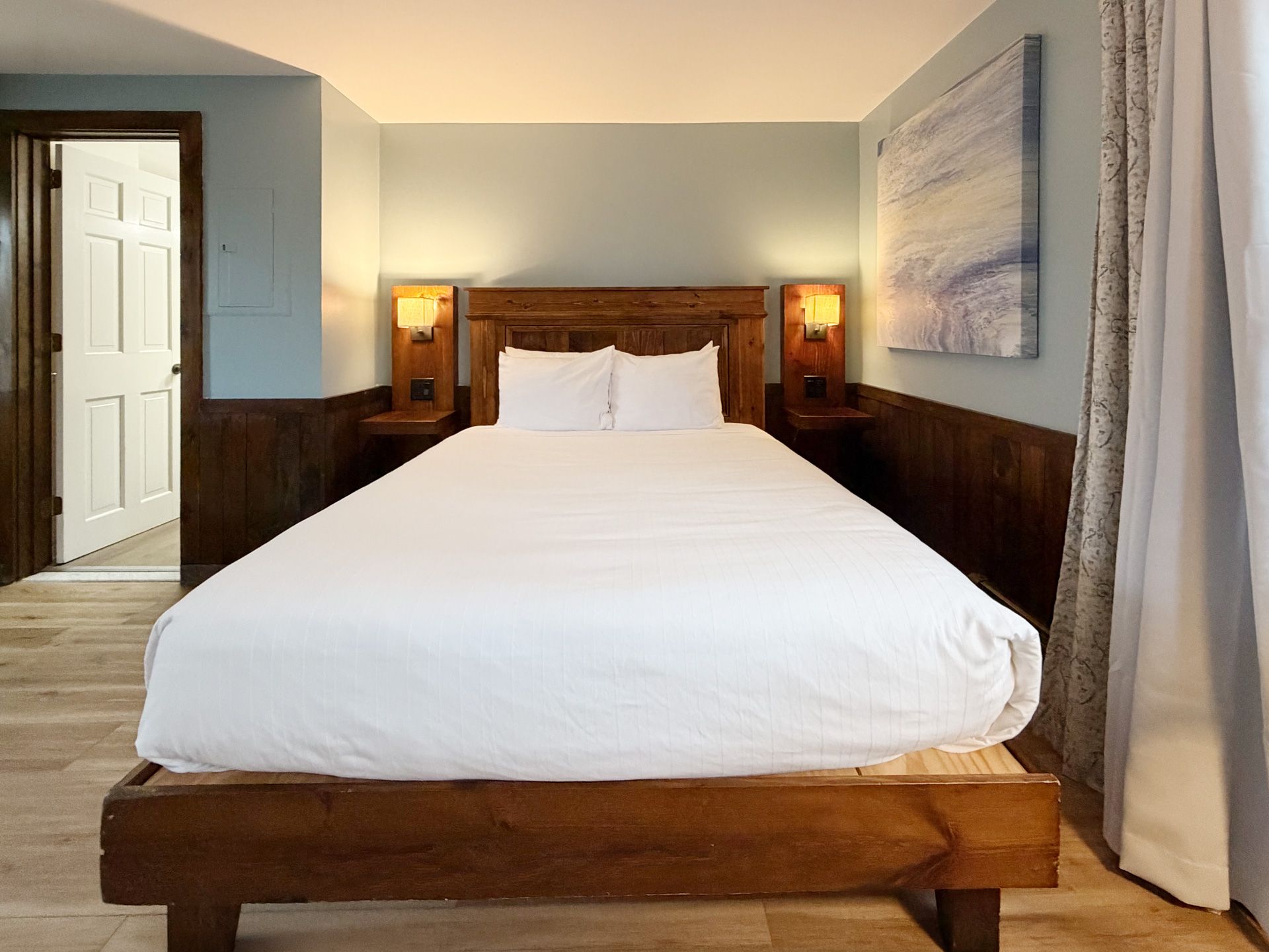 Neatly made bed in a cozy bedroom with wooden headboard, lamps, and a doorway on the left