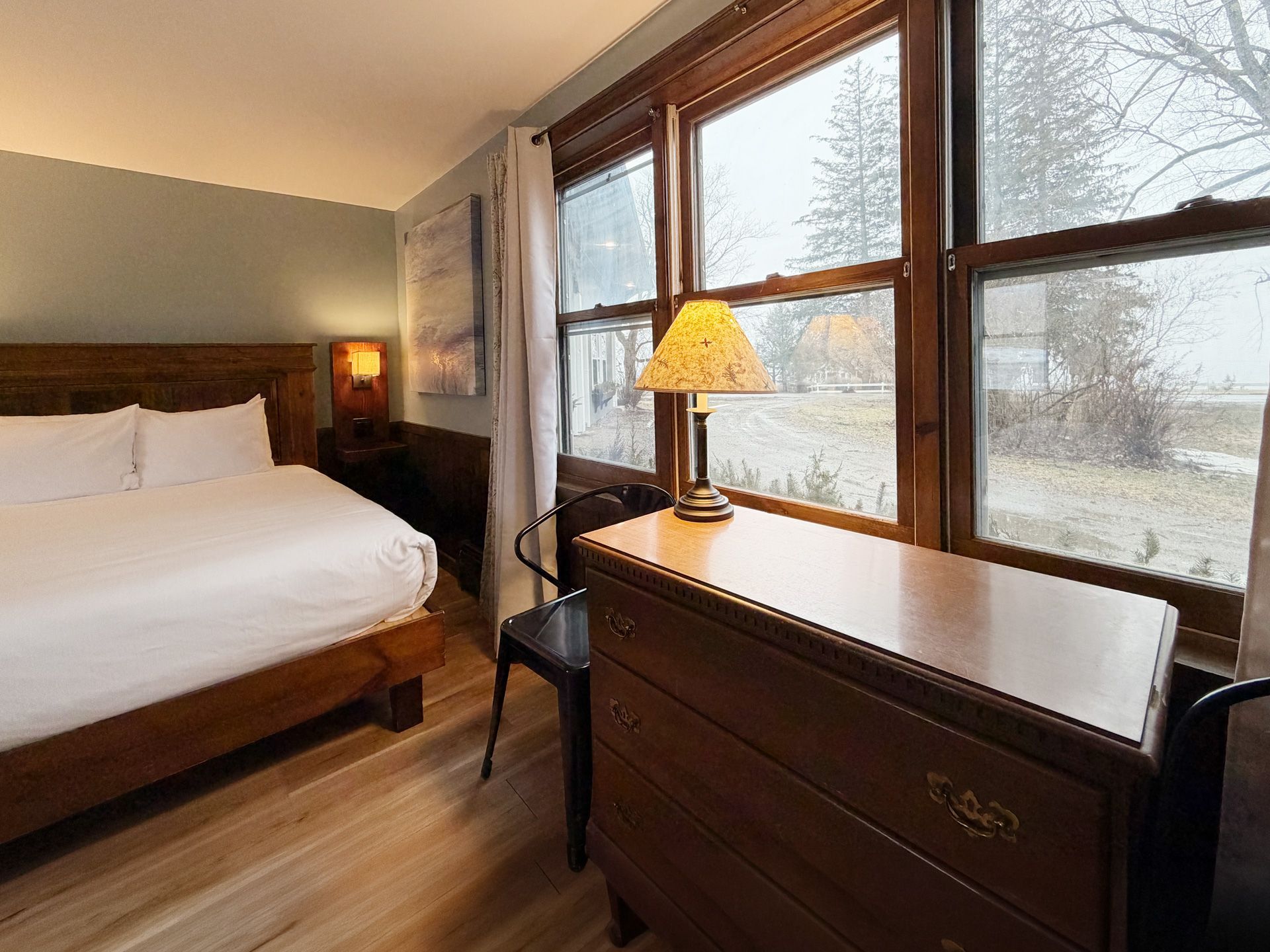 Cozy bedroom with white bed, wooden dresser, lamp, and large windows overlooking a snowy landscape
