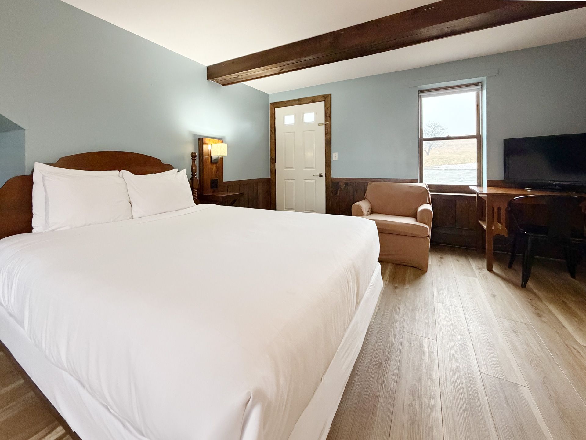 Bright bedroom with a large bed, armchair, desk, window, and wood ceiling beam