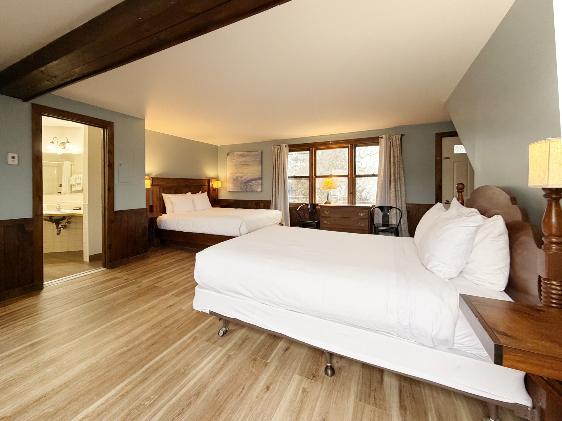 Spacious hotel room with two white beds, wood floors, and large windows with curtains