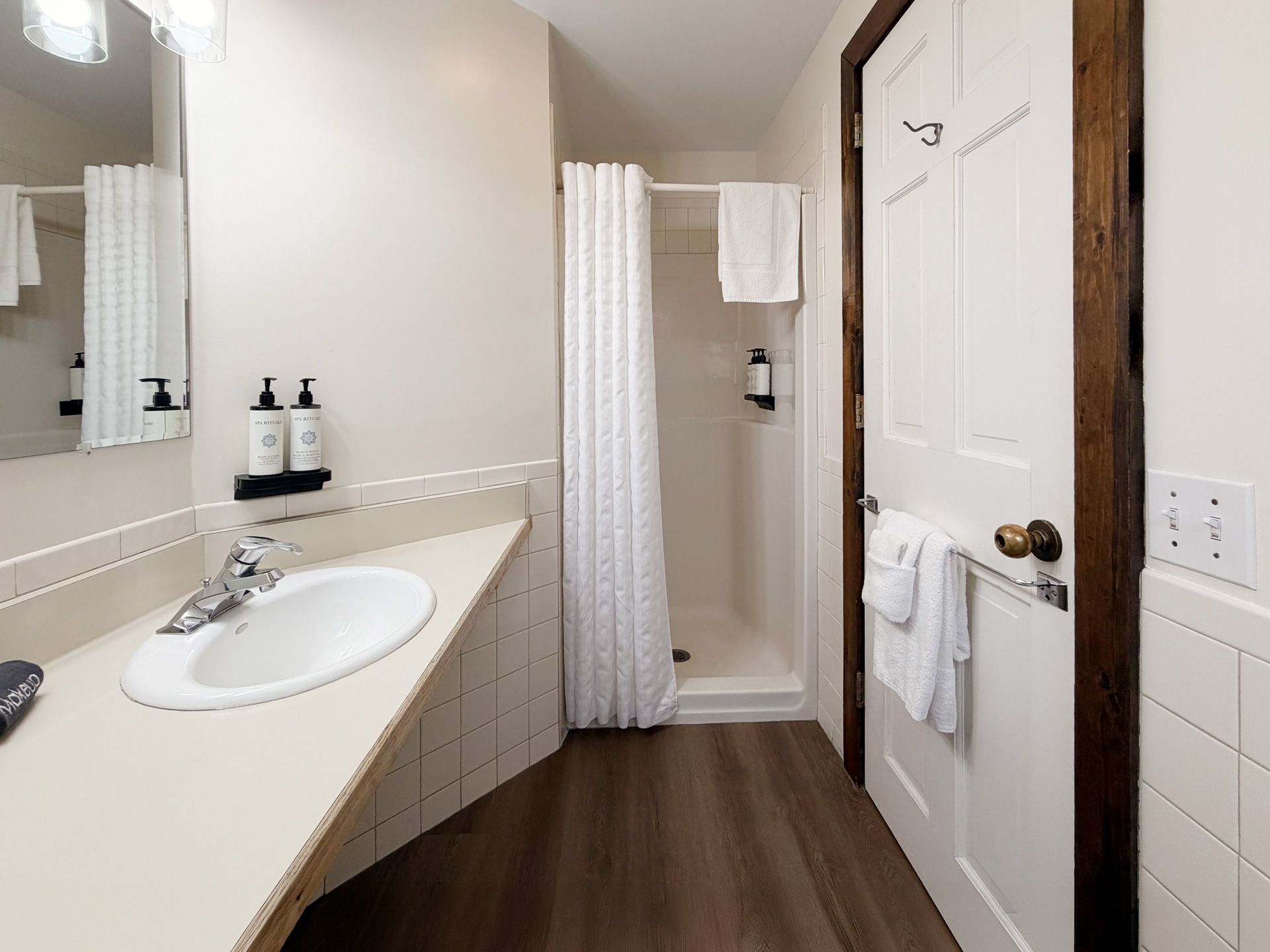 Small bathroom with vanity, mirror, shower curtain, and white door beside wood flooring.
