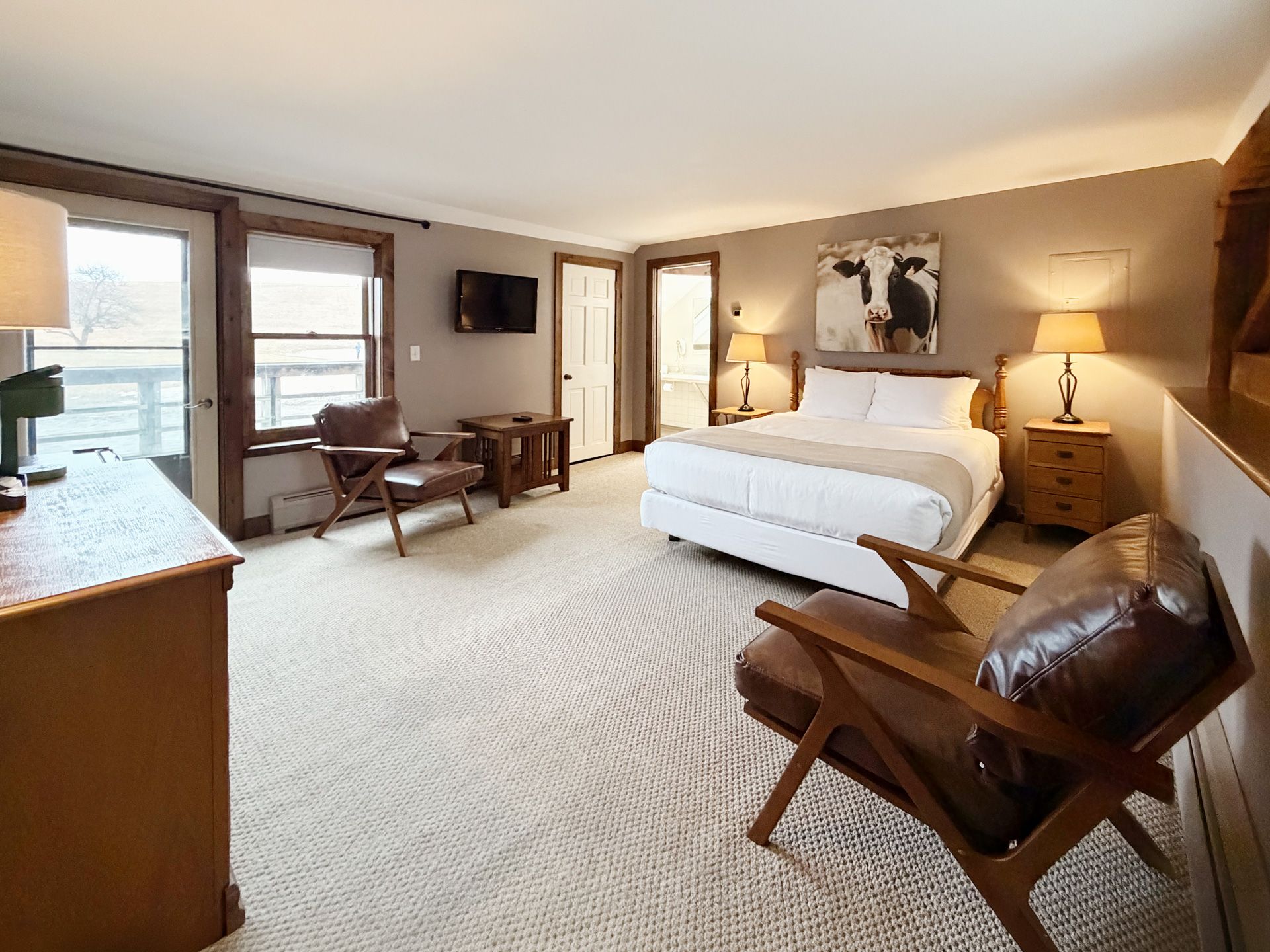 Bright bedroom with a white bed, wooden chairs, lamps, and large windows overlooking a calm view.