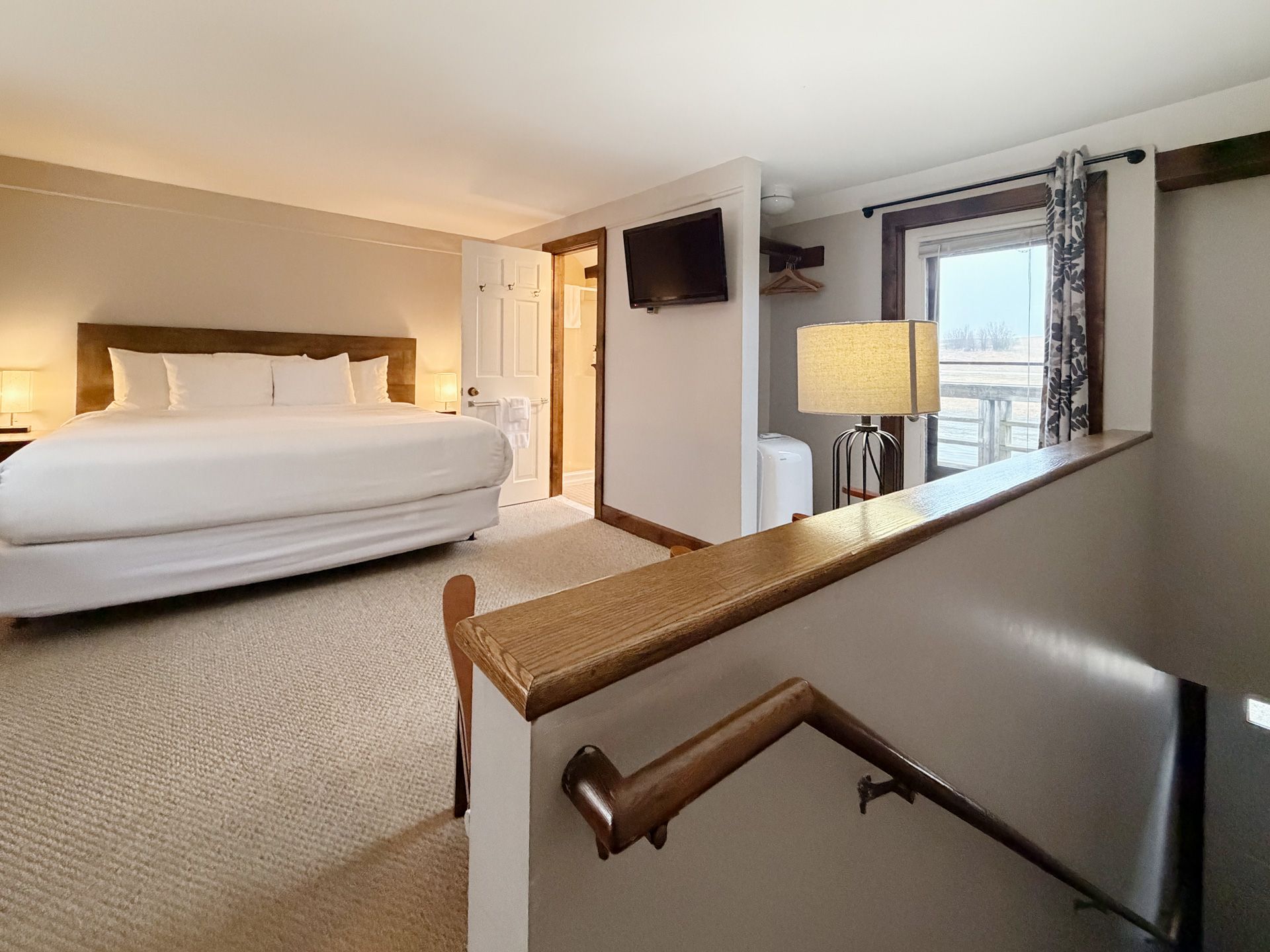 Bright hotel bedroom with king bed, carpeted floor, wall-mounted TV, and balcony view