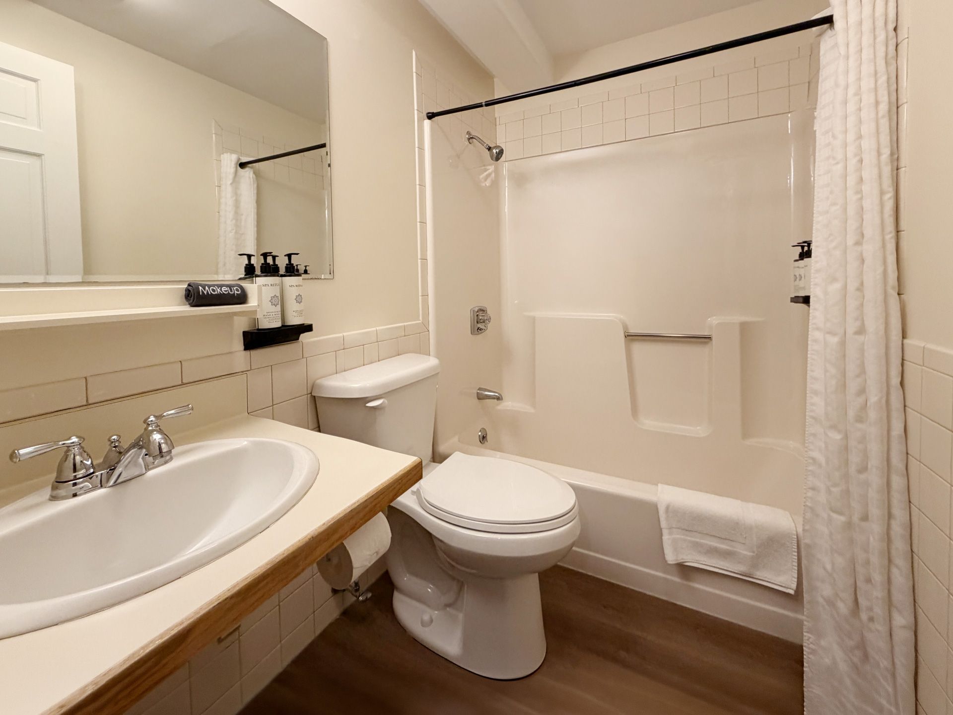 Small beige bathroom with sink, toilet, mirror, and shower/tub with white curtain and towel rack.