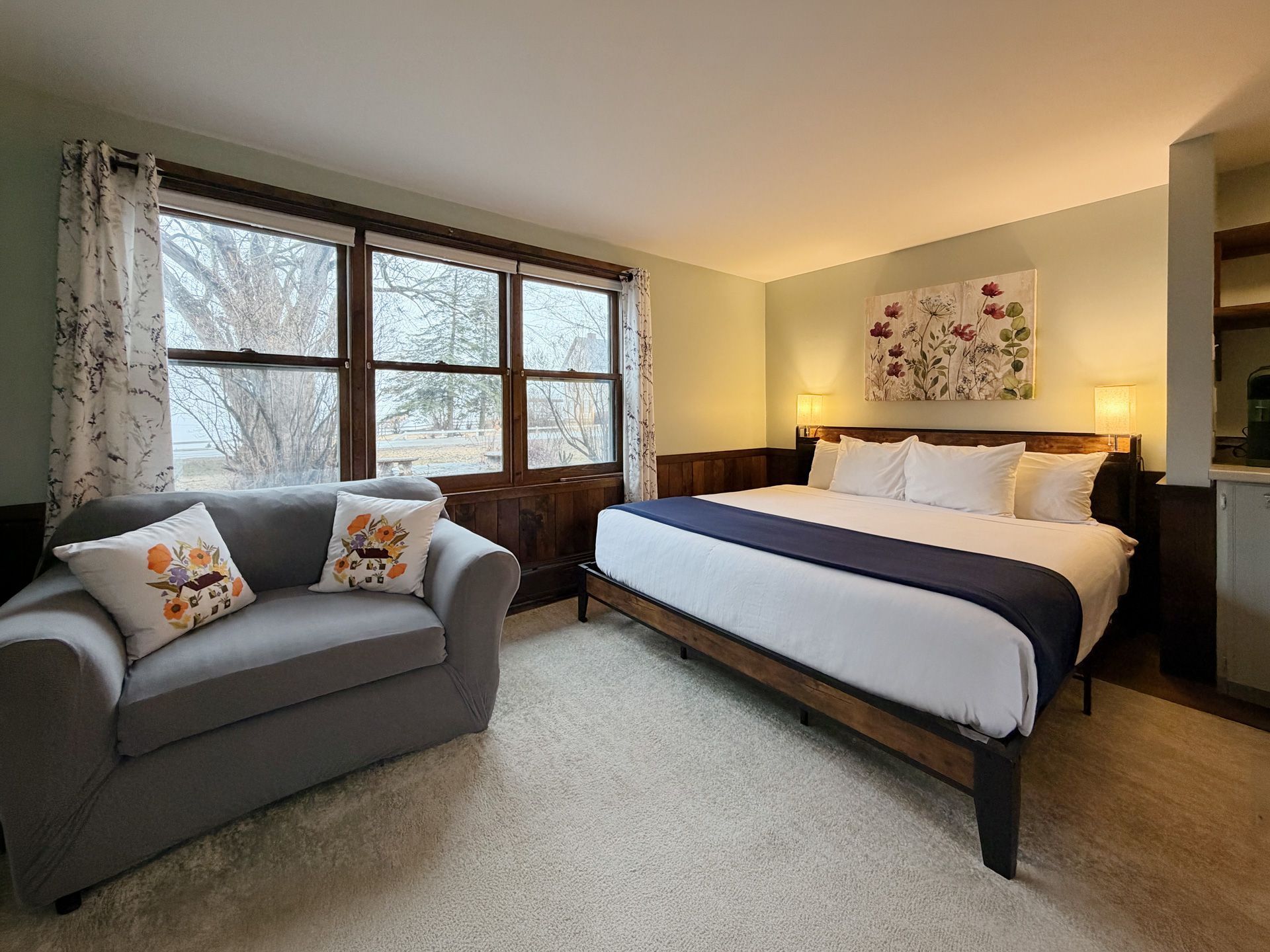 Cozy bedroom with a bed, gray loveseat, large window, warm lamps, and soft neutral decor