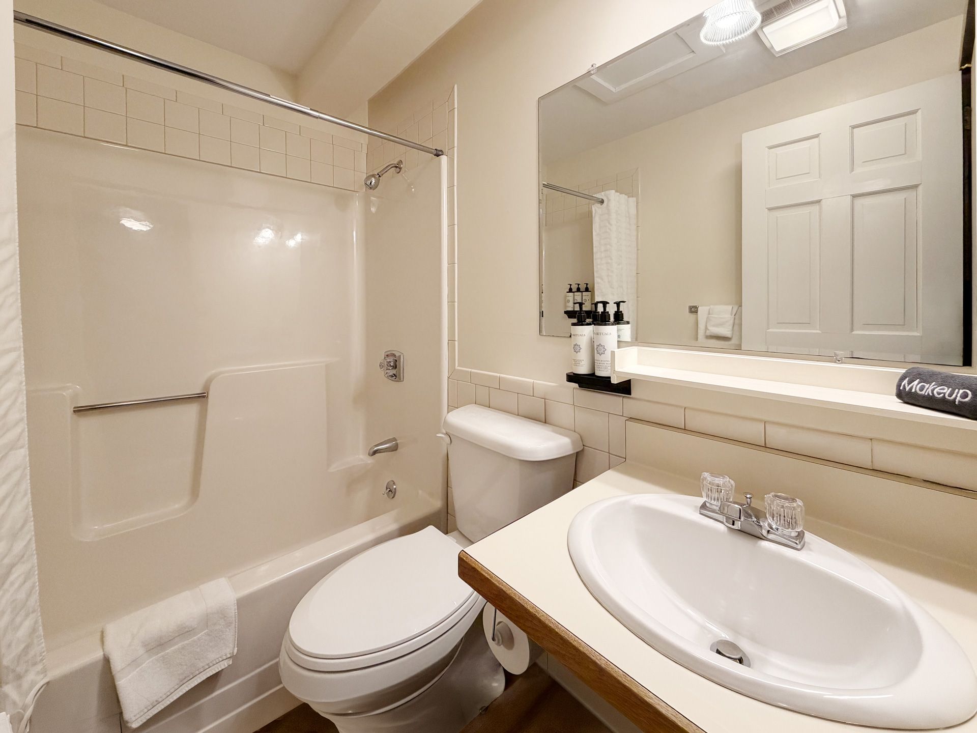 Compact beige bathroom with toilet, sink, bathtub, mirror, and white door.