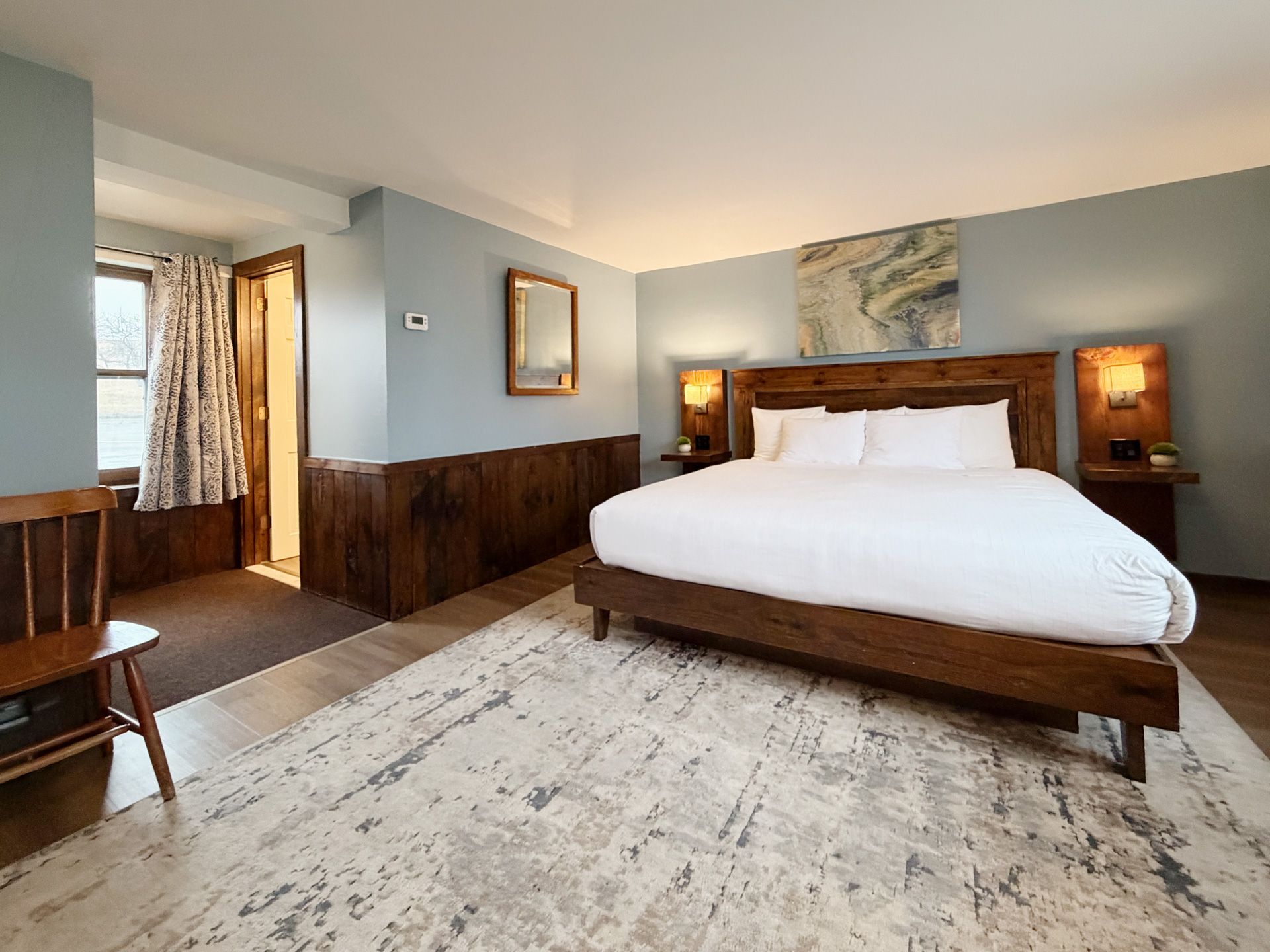 Spacious hotel bedroom with a large bed, wood furniture, pale blue walls, and a light patterned rug.