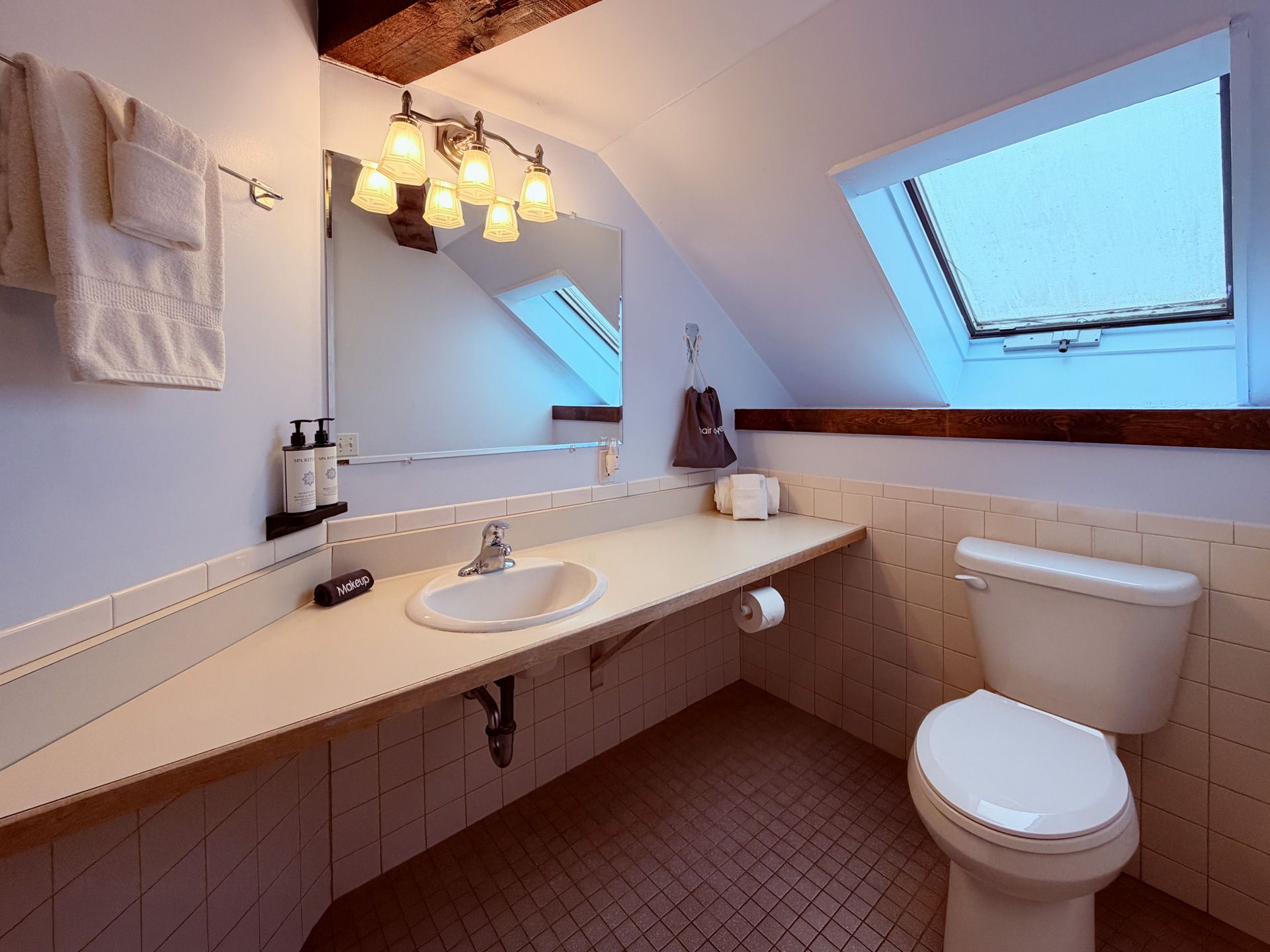Small attic bathroom with sloped skylights, beige tile, double sink, and toilet.