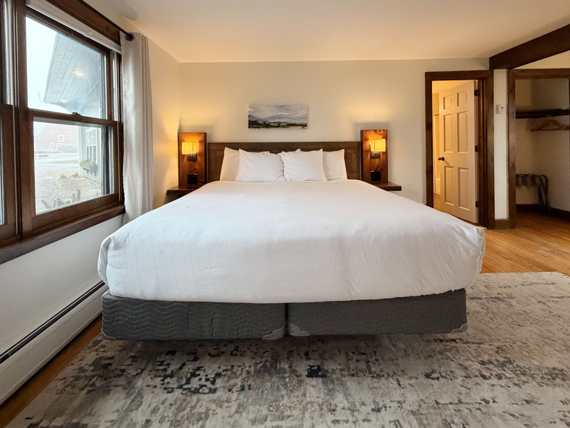 Bright bedroom with a large white bed, bedside lamps, window, and open door to a hallway
