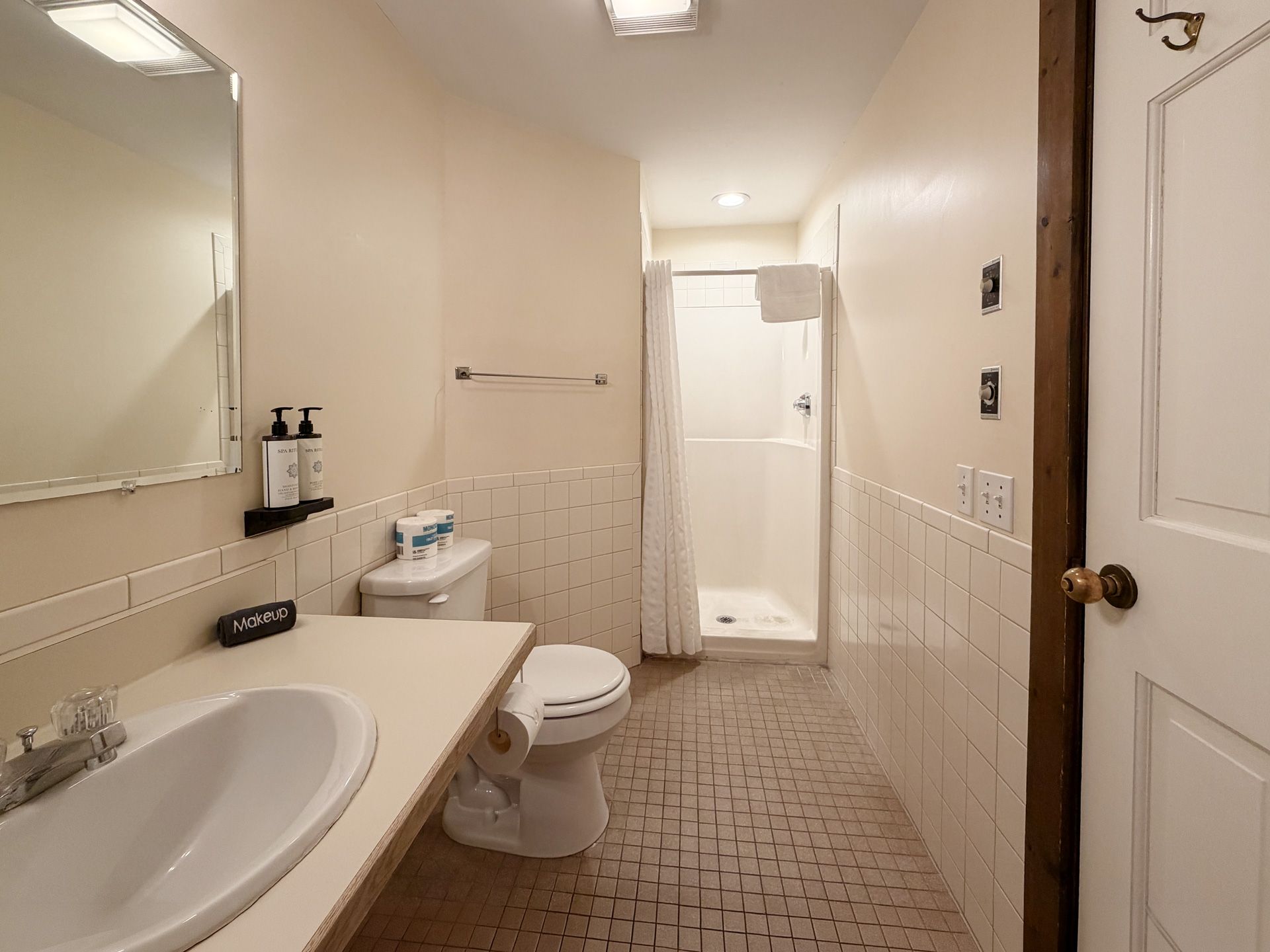 Bright bathroom with sink, toilet, mirror, shower, and tiled floor; beige walls and white fixtures.
