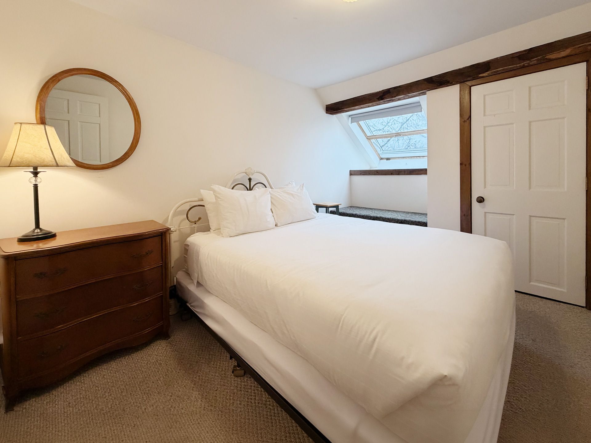 Small bedroom with a white bed, wooden dresser, round mirror, and window seat under a slanted ceiling