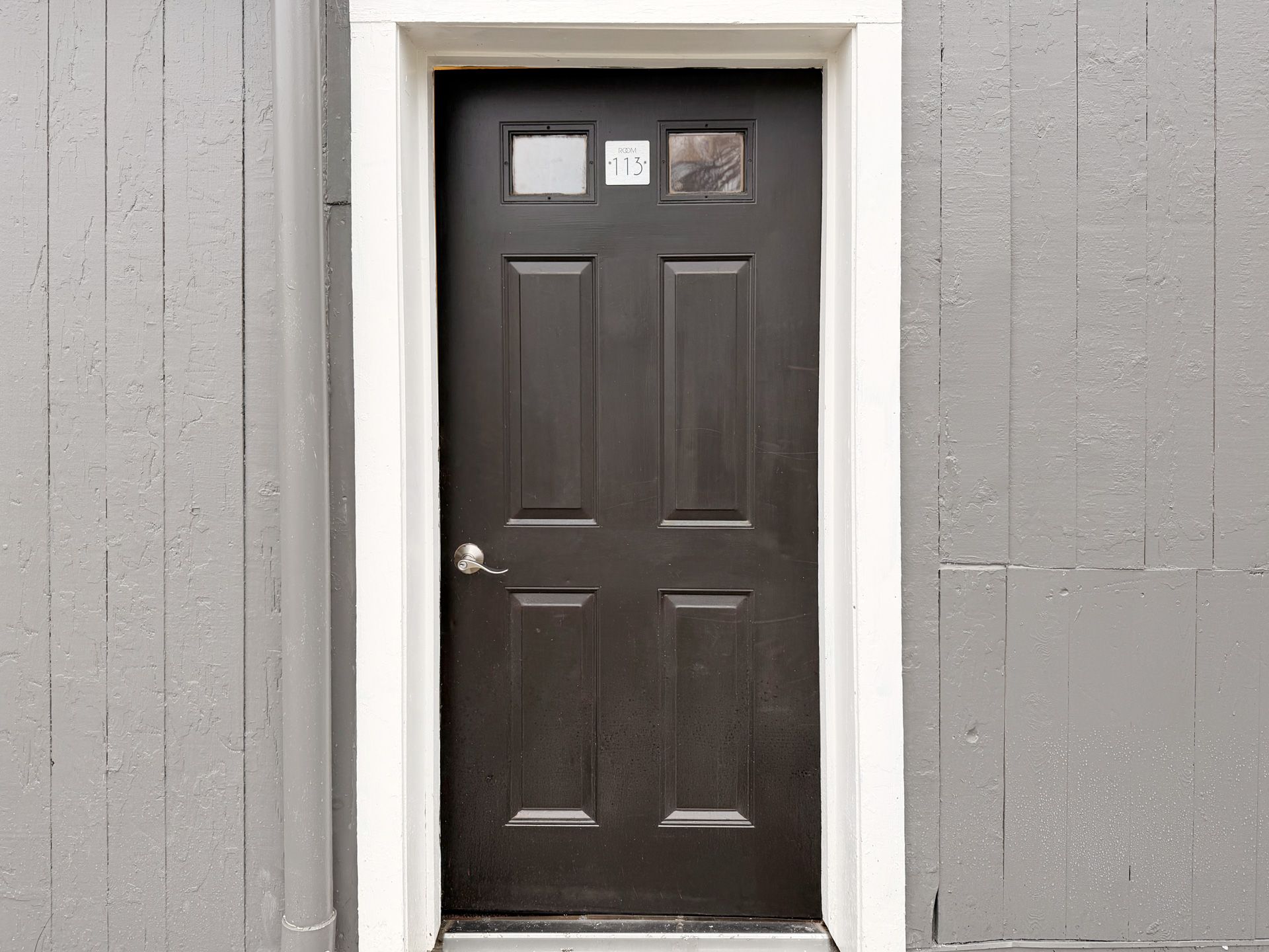 Dark brown front door with small windows, set in a white frame on a gray exterior wall.