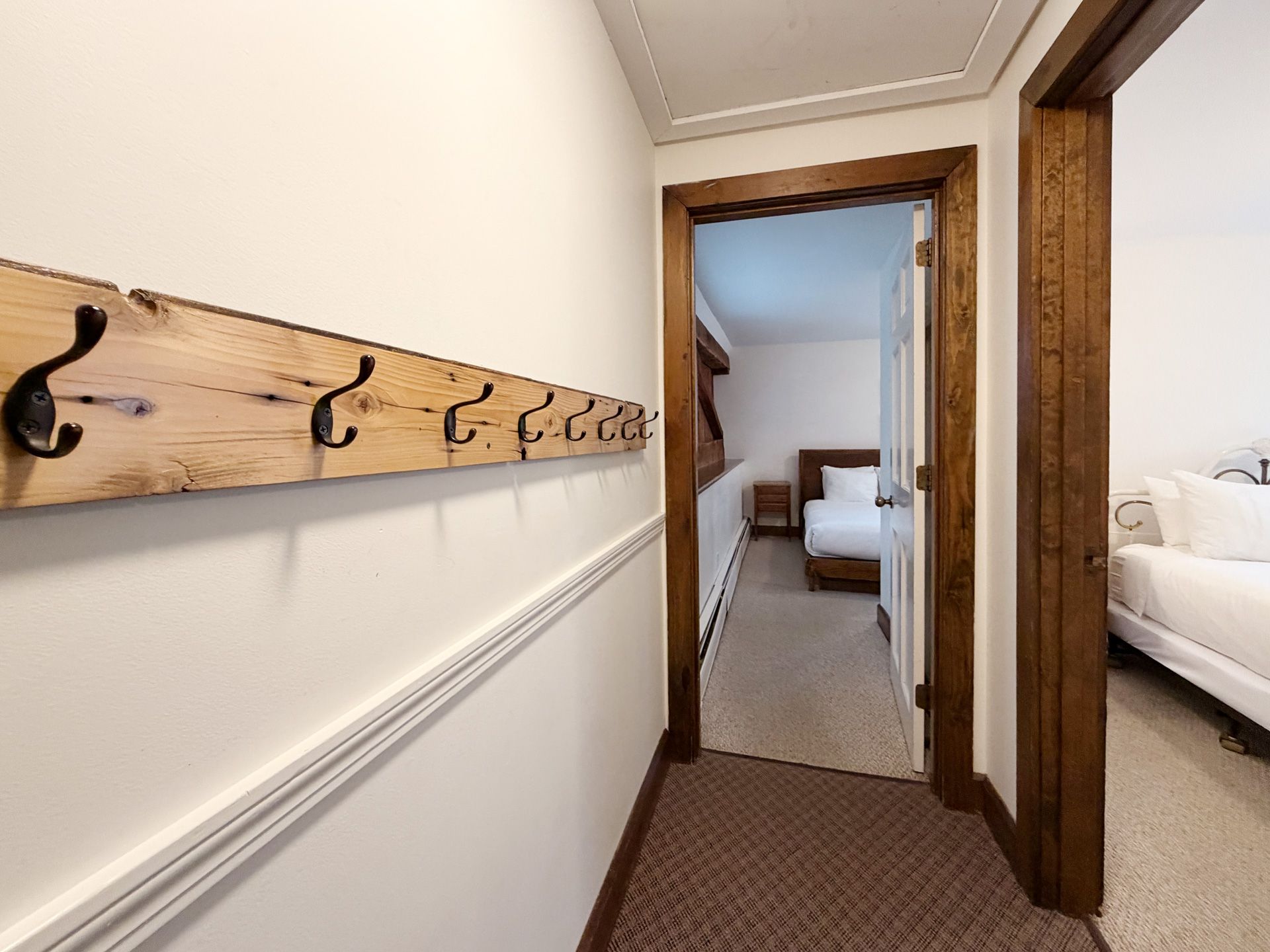 Narrow hallway with wooden coat rack leading to a bedroom with a bed and carpeted floor.