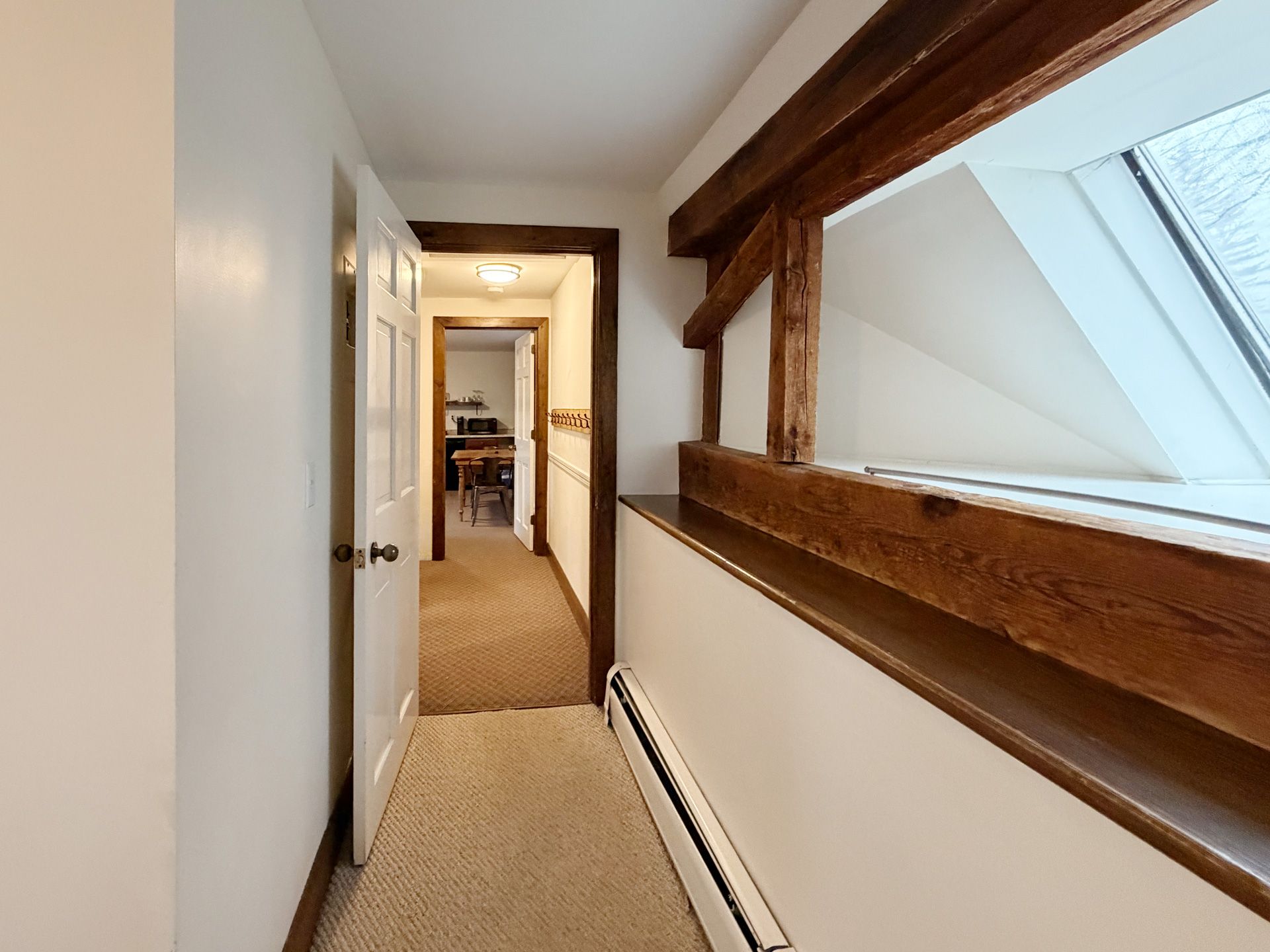 Narrow carpeted hallway with white walls, wooden beam, and skylight above stairs.