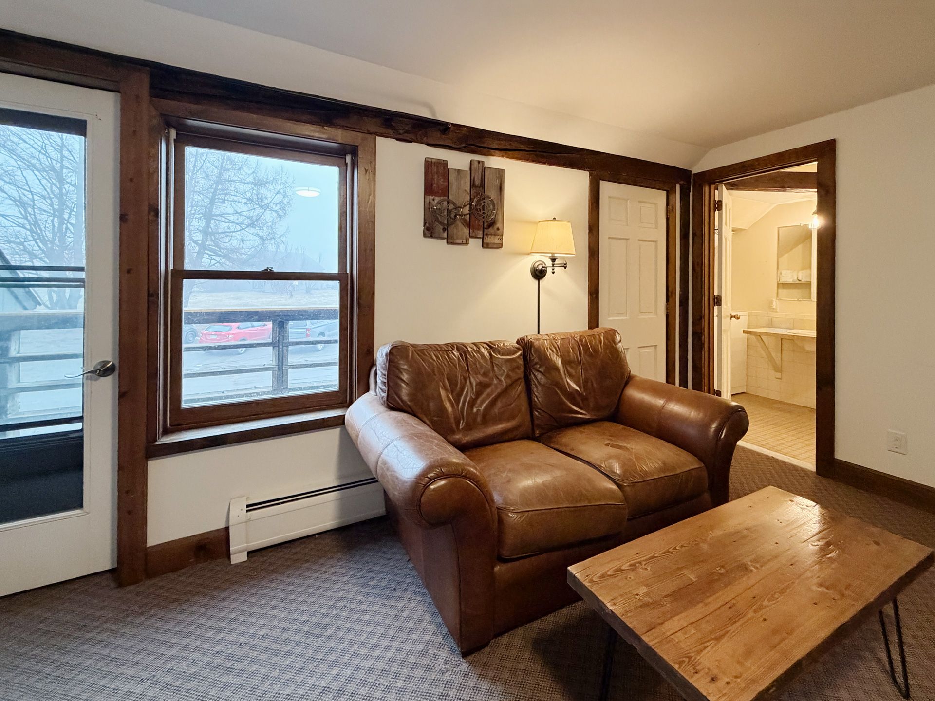 Cozy living room with brown leather couch, wooden coffee table, large windows, and adjacent bathroom doorway