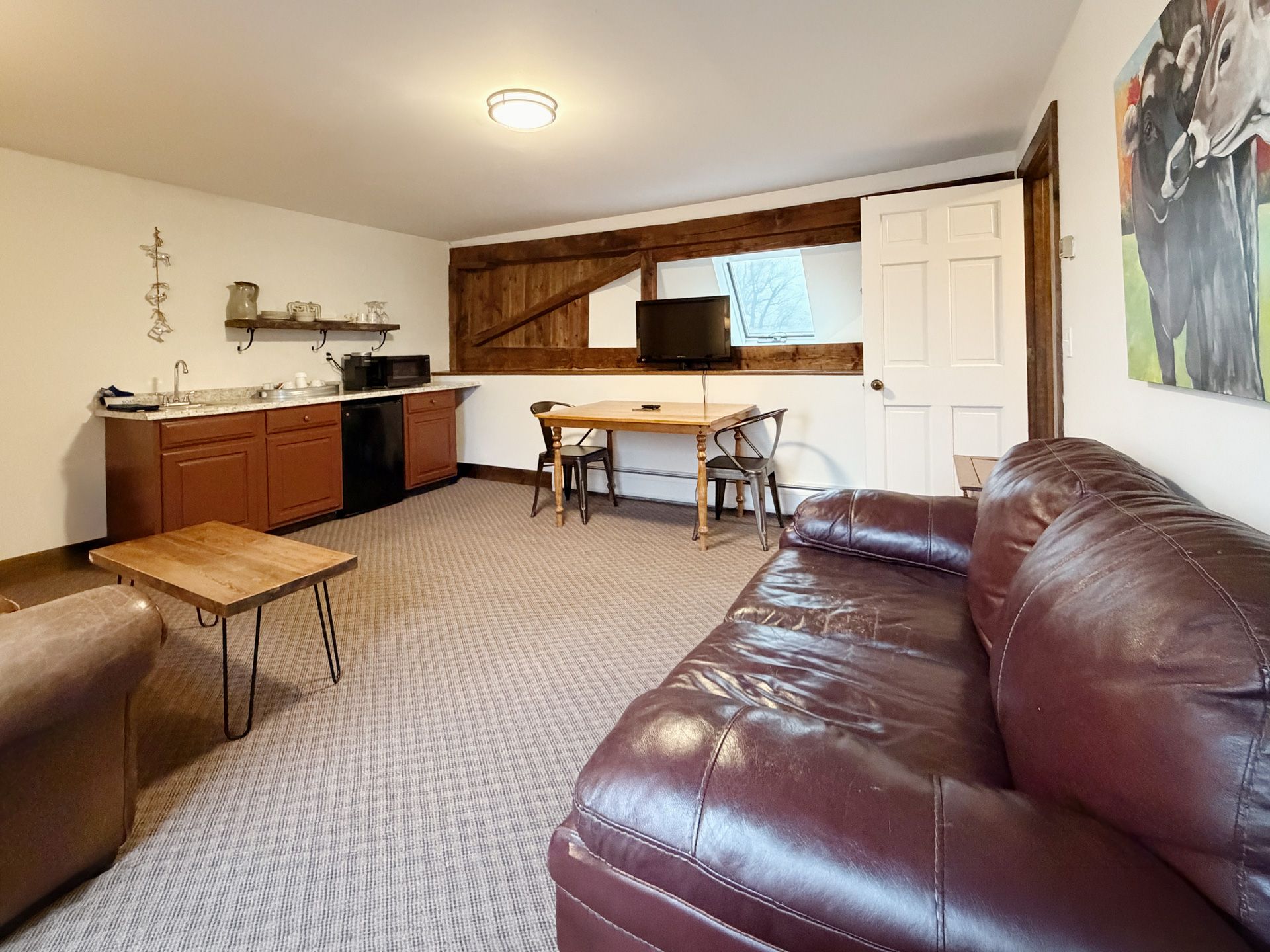 Living room with brown leather sofa, small tables, kitchen area, and TV in the background